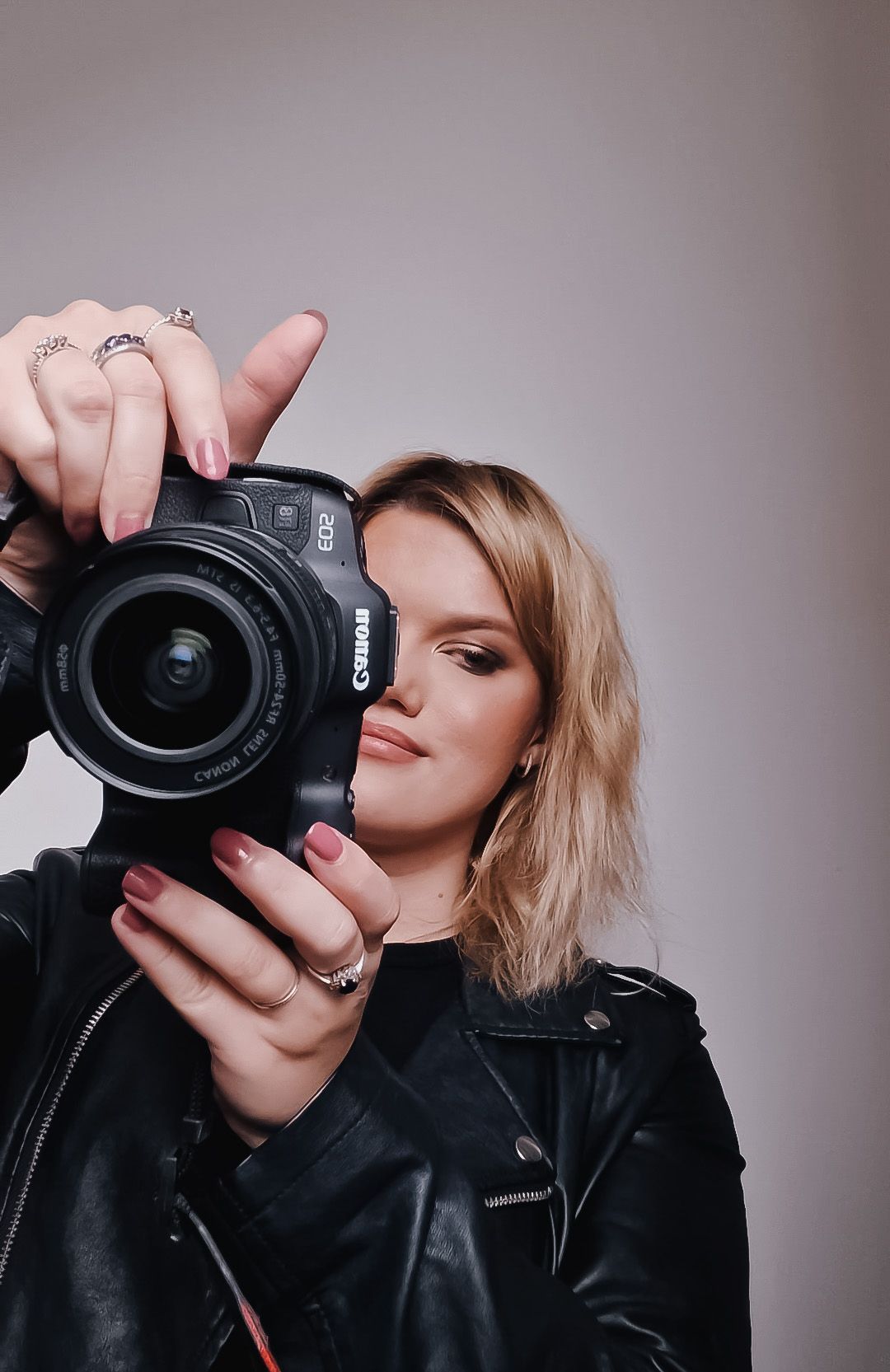 Young woman in leather jacket holding a Canon DSLR camera up to her face, with rings and pink nails.
