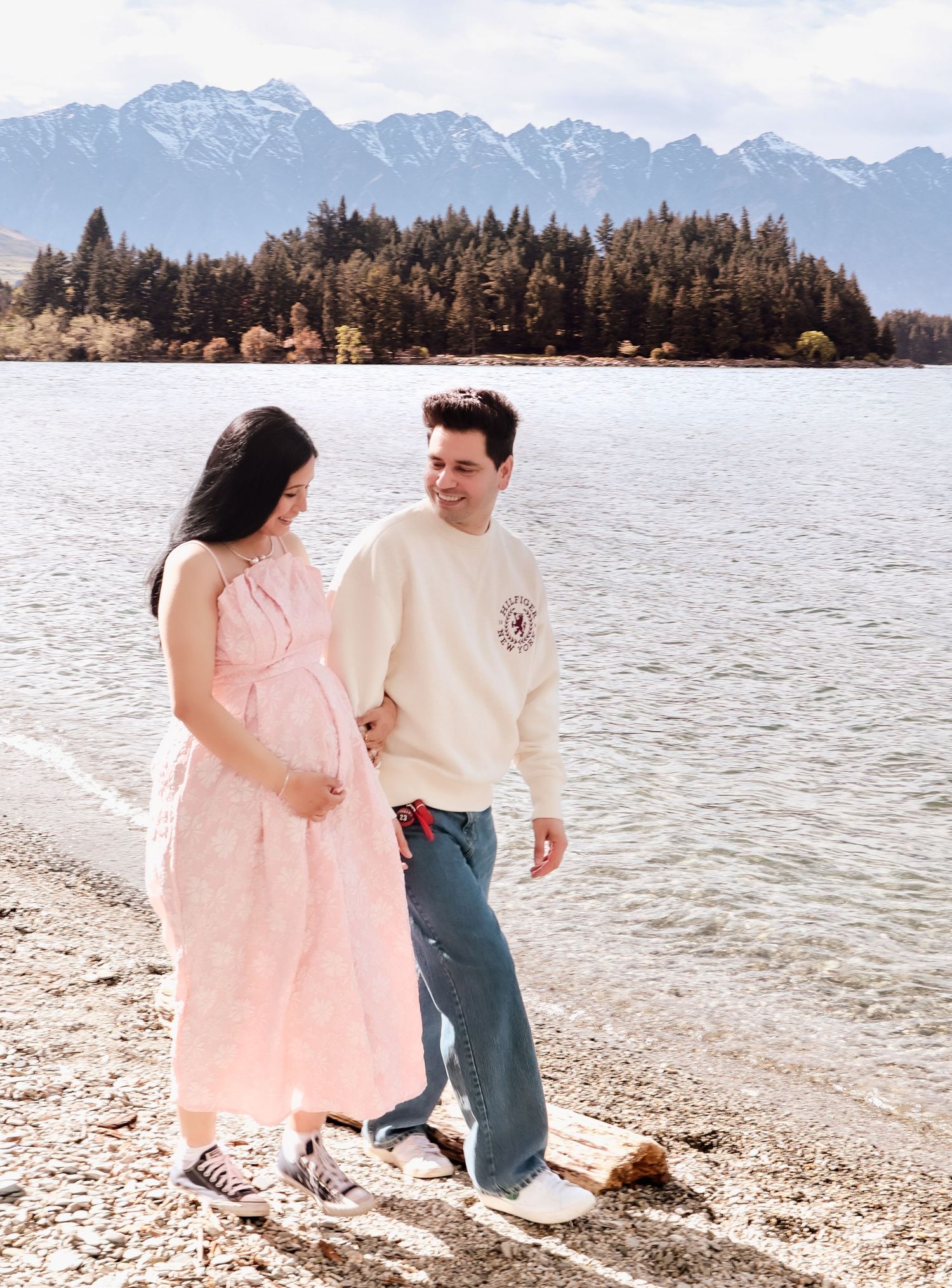 Pregnant woman in pink dress walks with partner along a lakeside with snowy mountain backdrop.