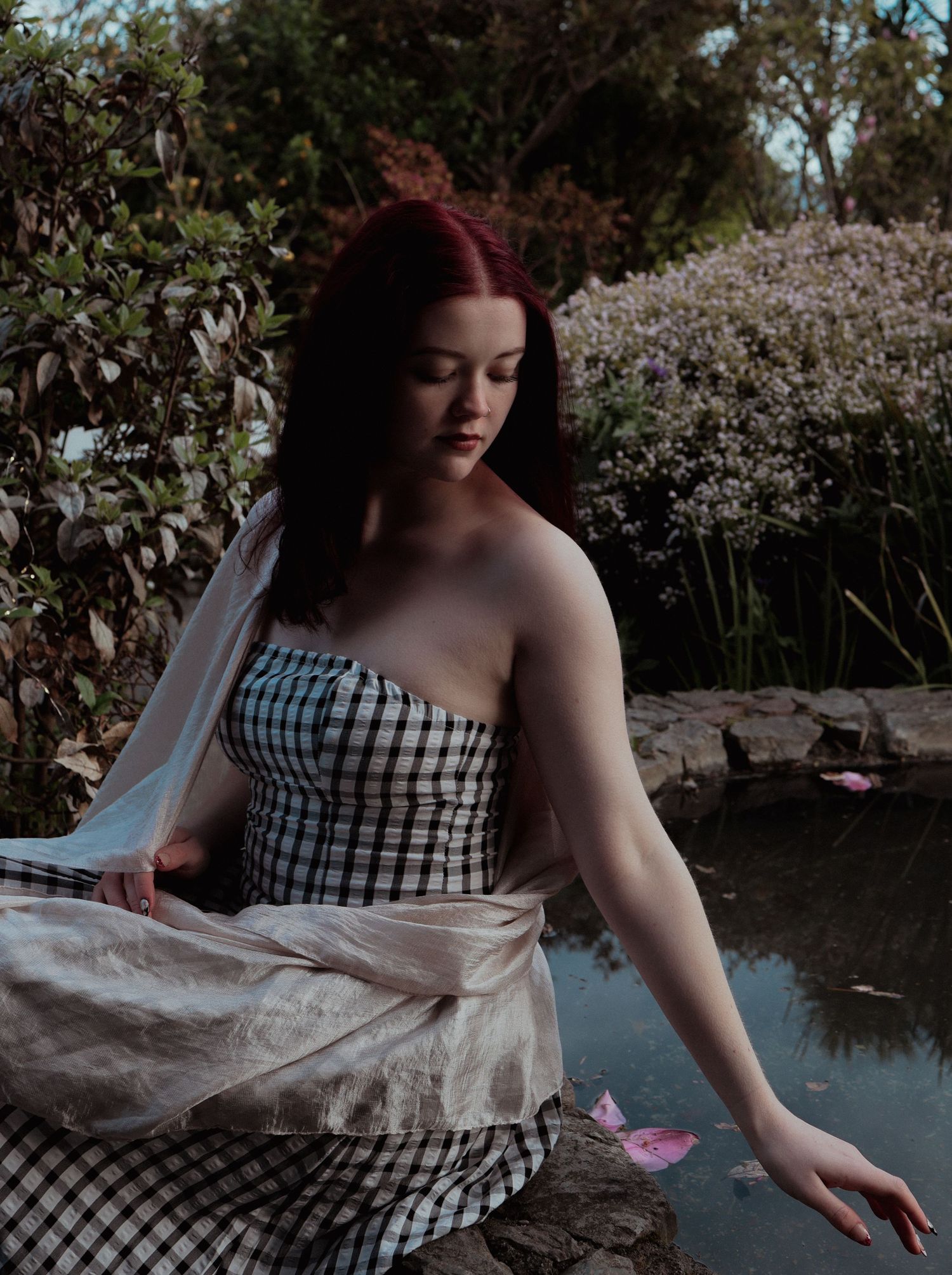 A woman with red hair in a checkered strapless dress sits gracefully beside a garden pond.