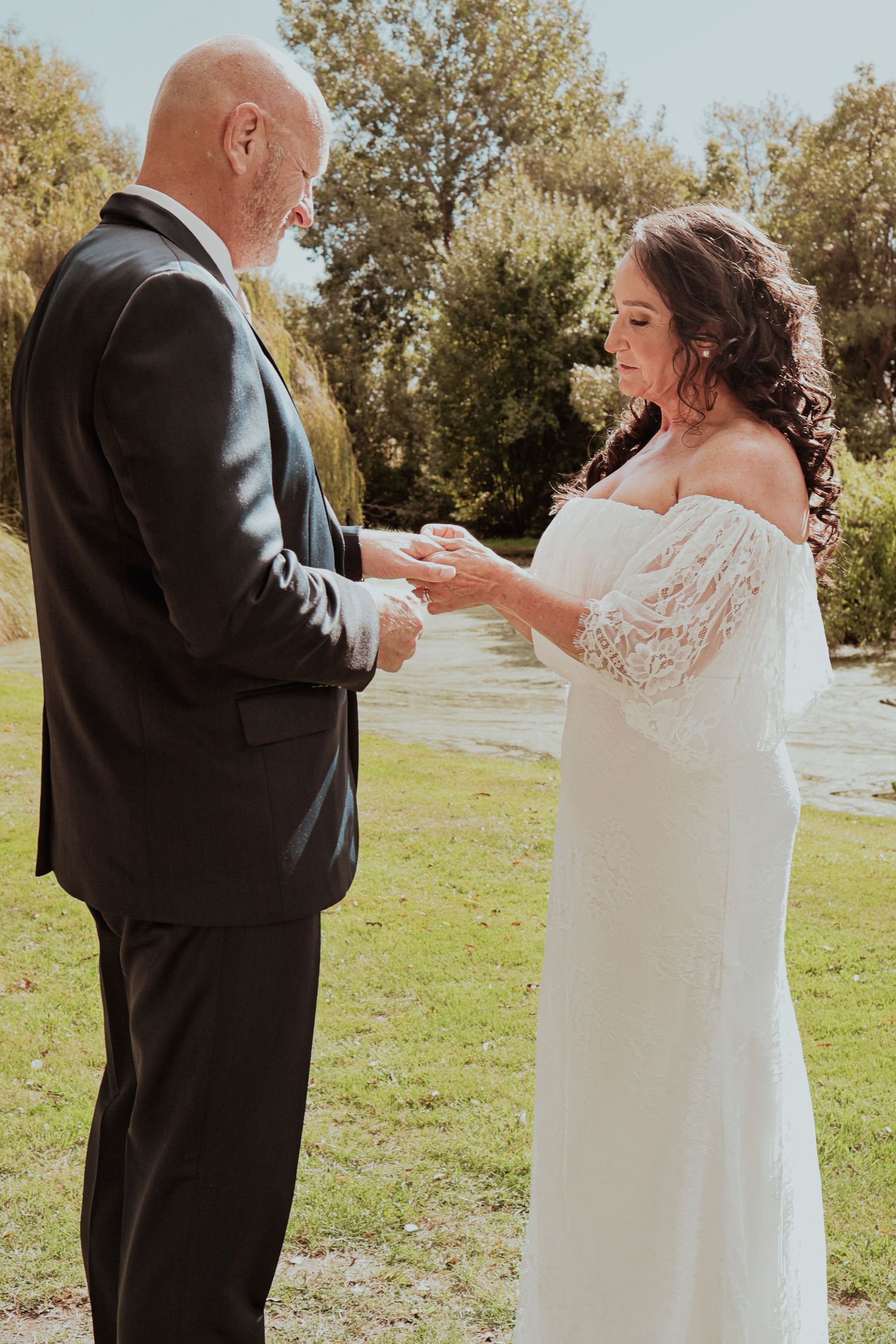 Groom places wedding ring on bride's finger during outdoor ceremony beside tranquil river.