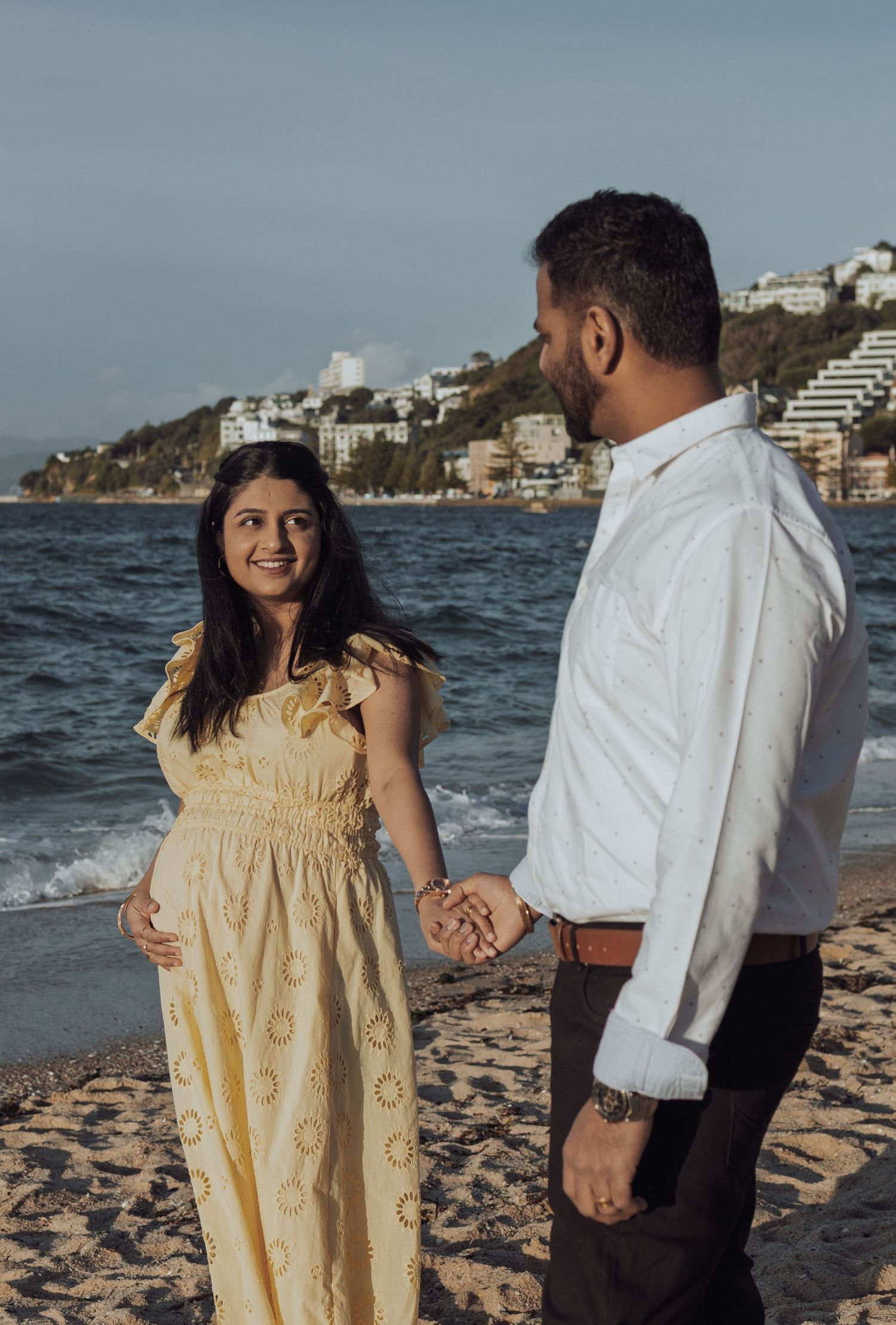 Pregnant woman in yellow dress holds hands with man in white shirt on sandy beach with ocean backdrop.