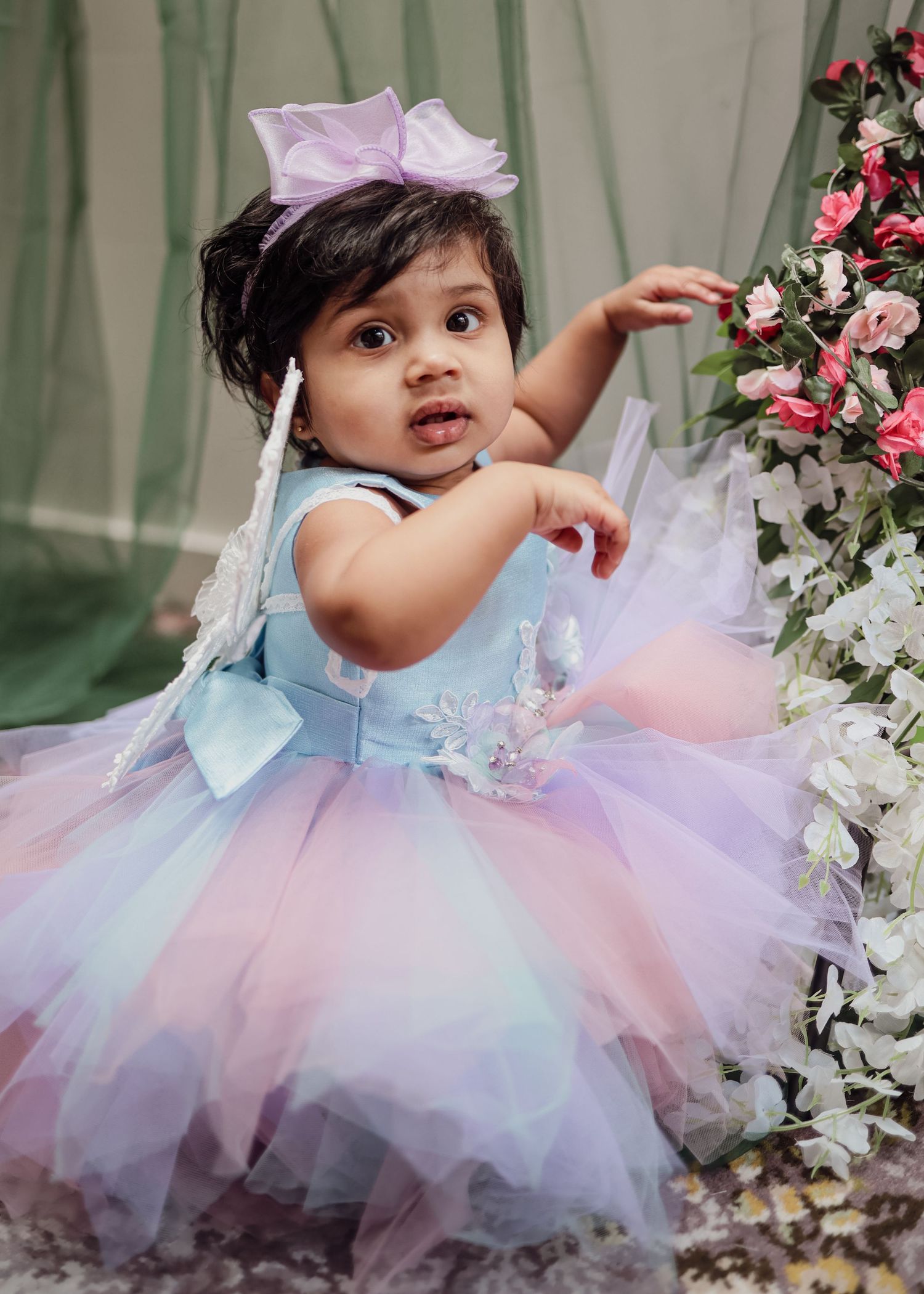 Adorable baby girl in pastel rainbow tutu dress with purple bow headband near floral decorations.