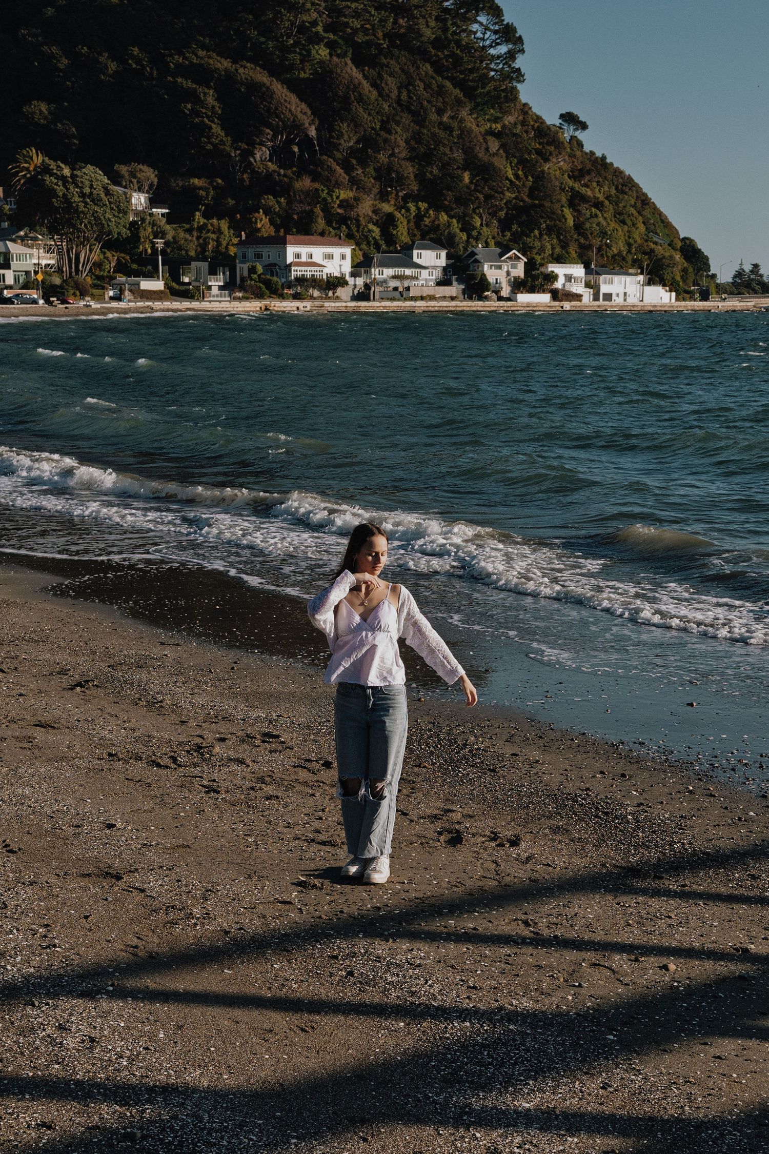 Woman walking on a sandy beach with ocean waves, green hillside, and white houses in the background.