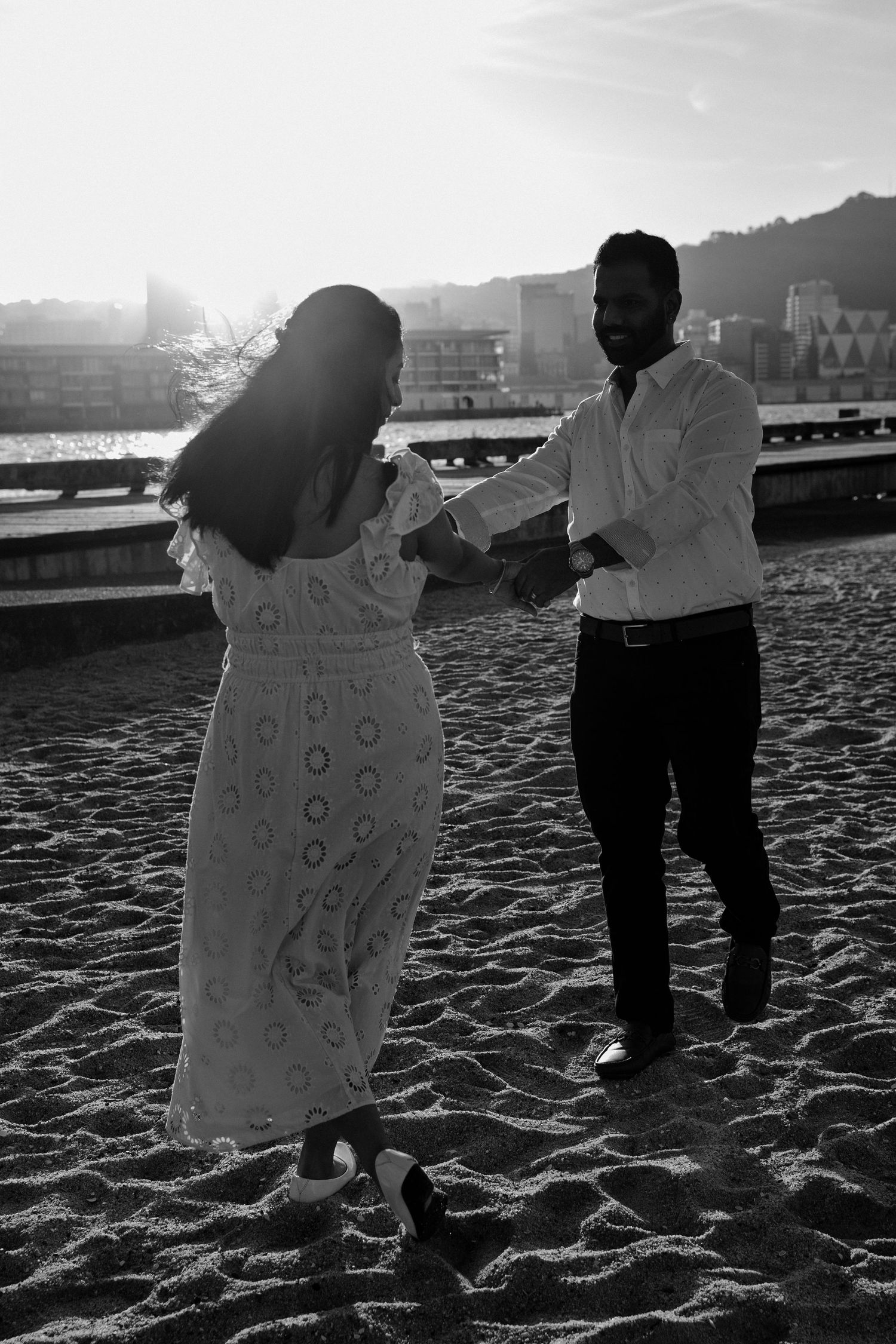 Couple dancing on sandy beach at sunset in black and white photo, woman in white dress, man in white shirt.