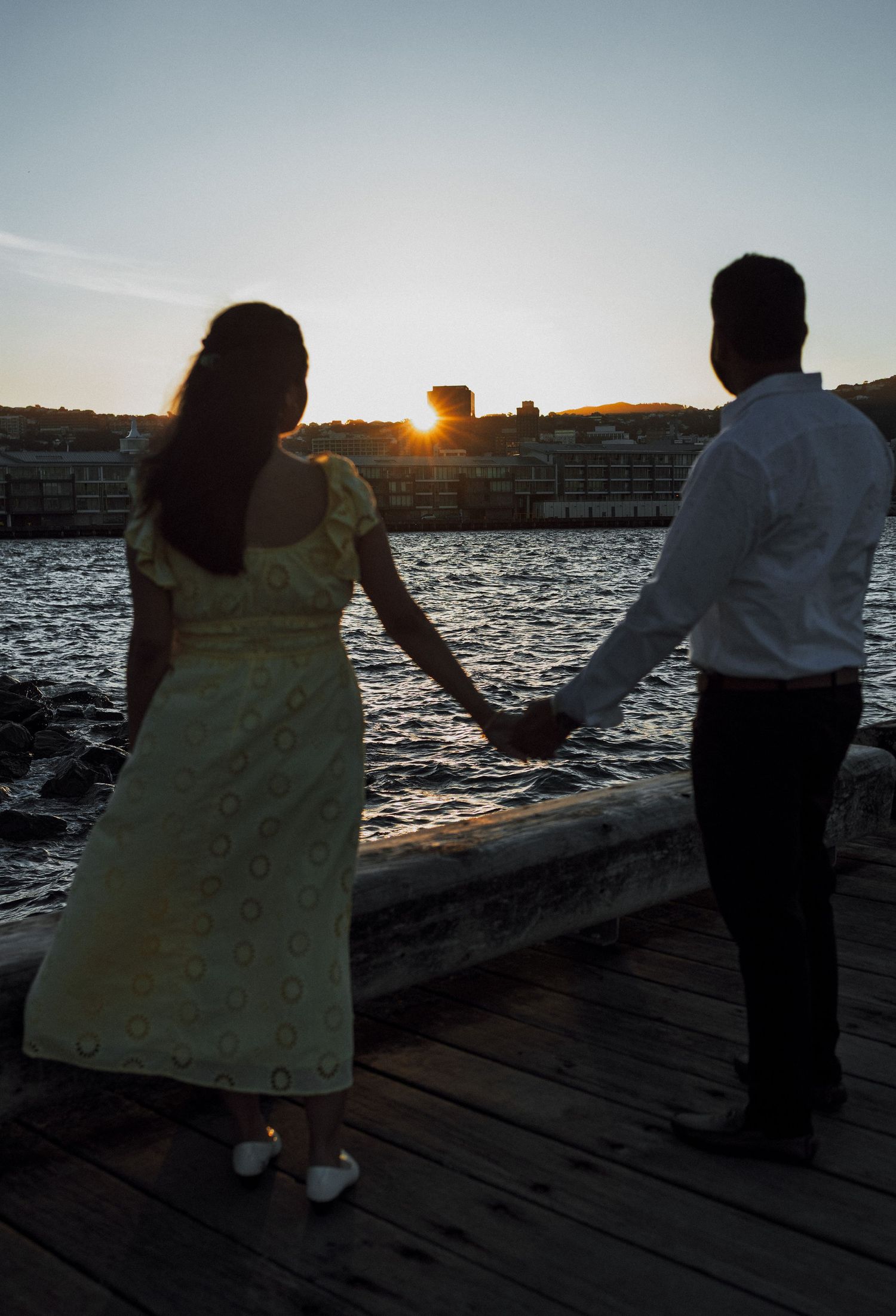 Couple holding hands on a wooden pier at sunset with waterfront buildings in the background.