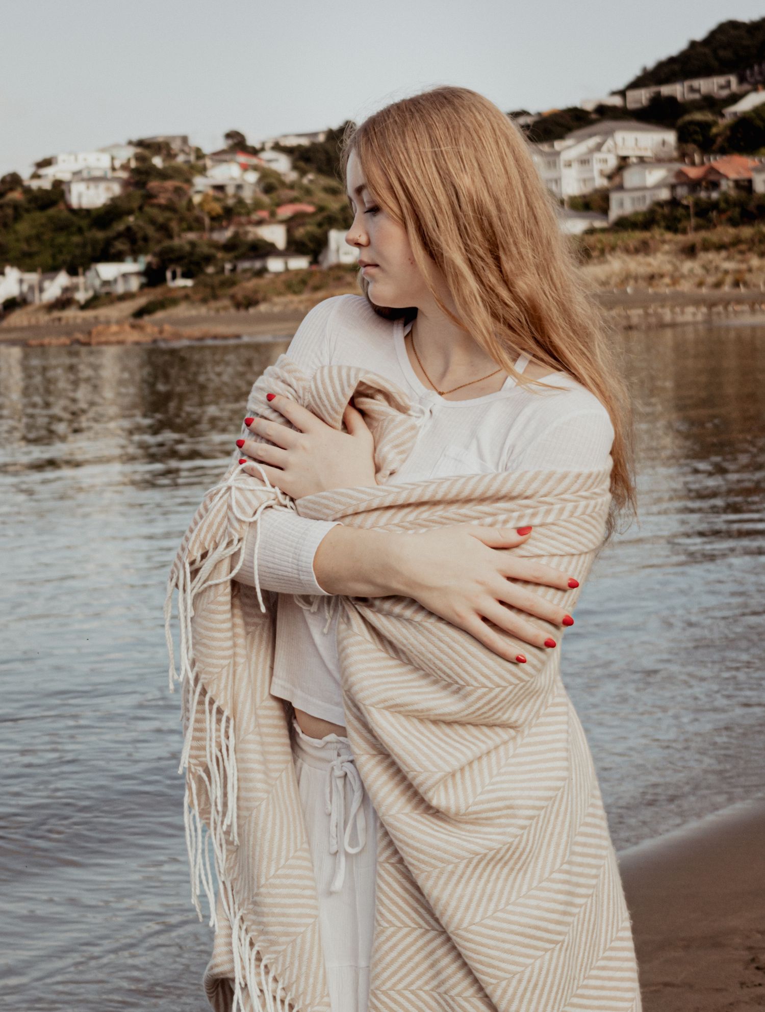 Blonde woman wrapped in beige striped blanket standing by a calm coastal bay.
