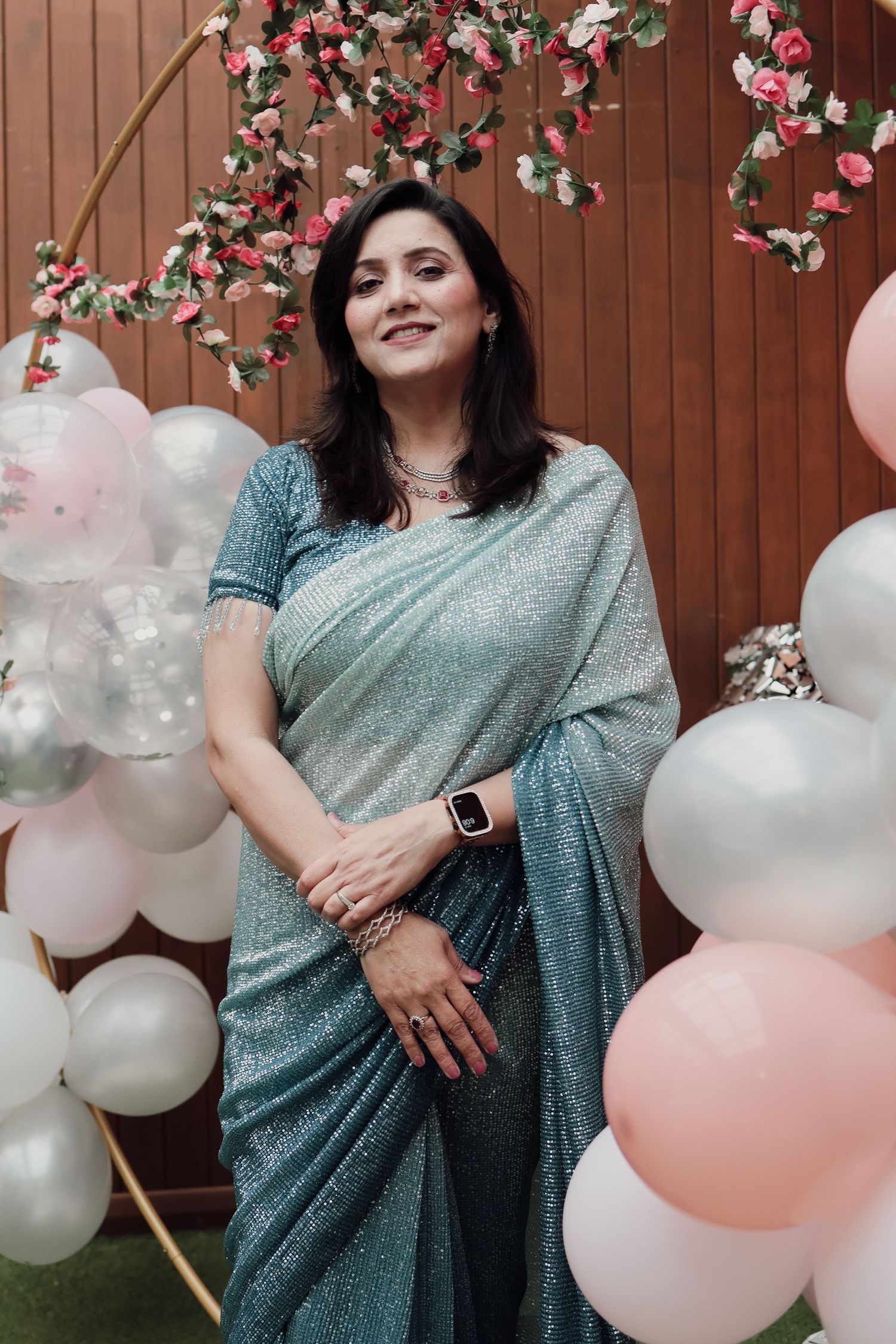 Woman smiling in a blue sparkly saree surrounded by pink and white balloons at a party.