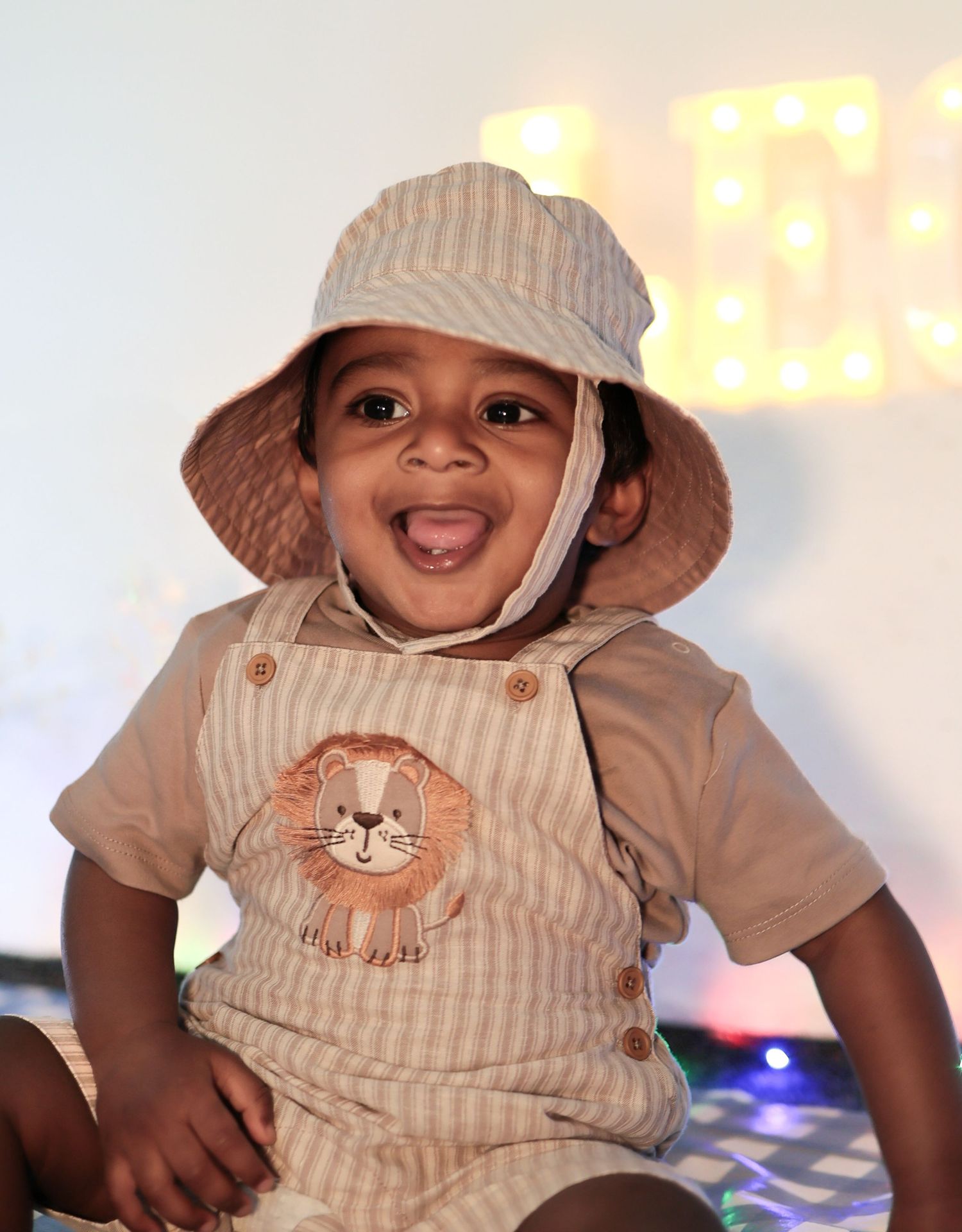 Adorable toddler in striped overalls and bucket hat smiles joyfully at birthday celebration.