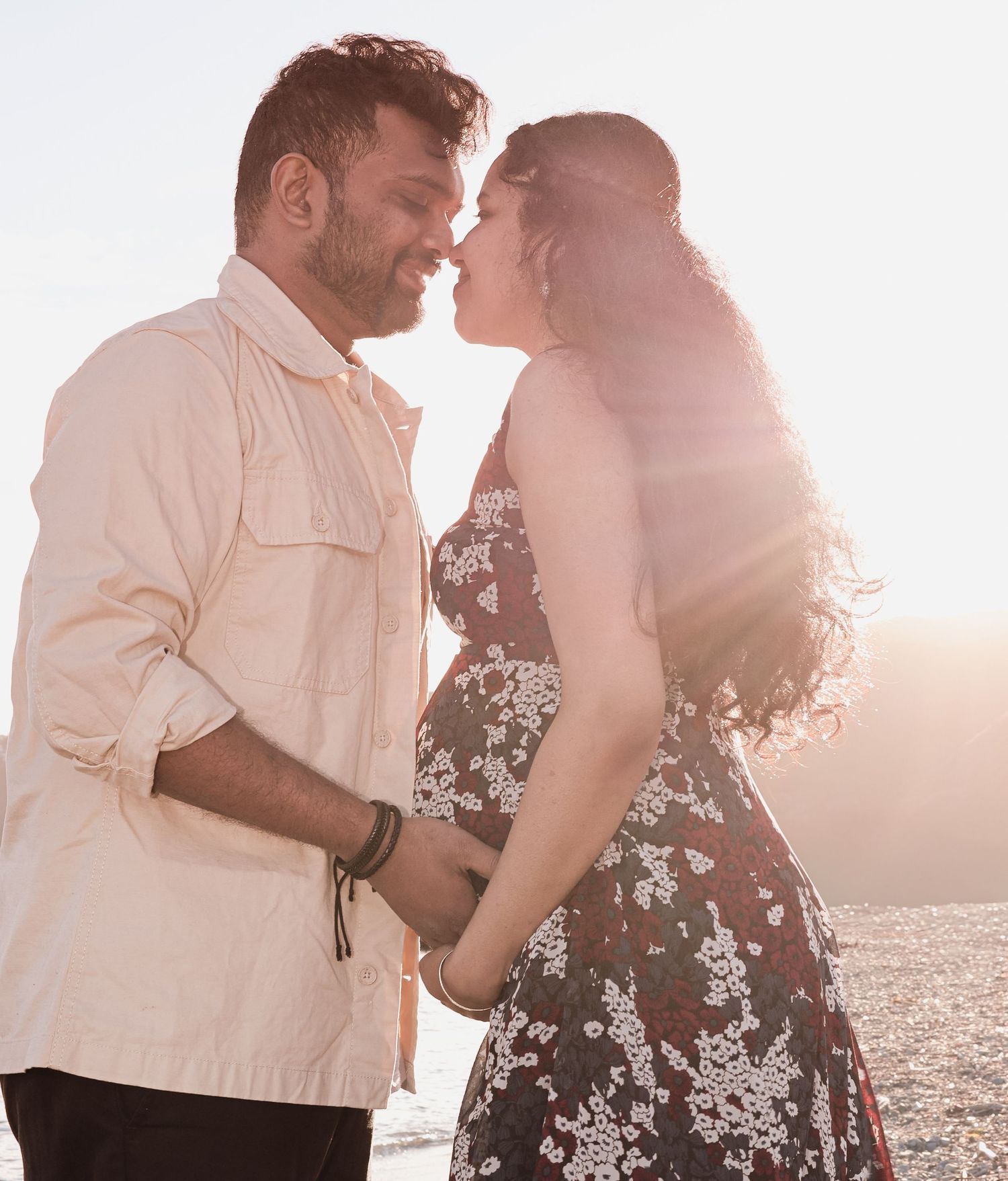 Expecting couple nearly kissing in golden backlit sunlight, woman in floral dress holds baby bump.