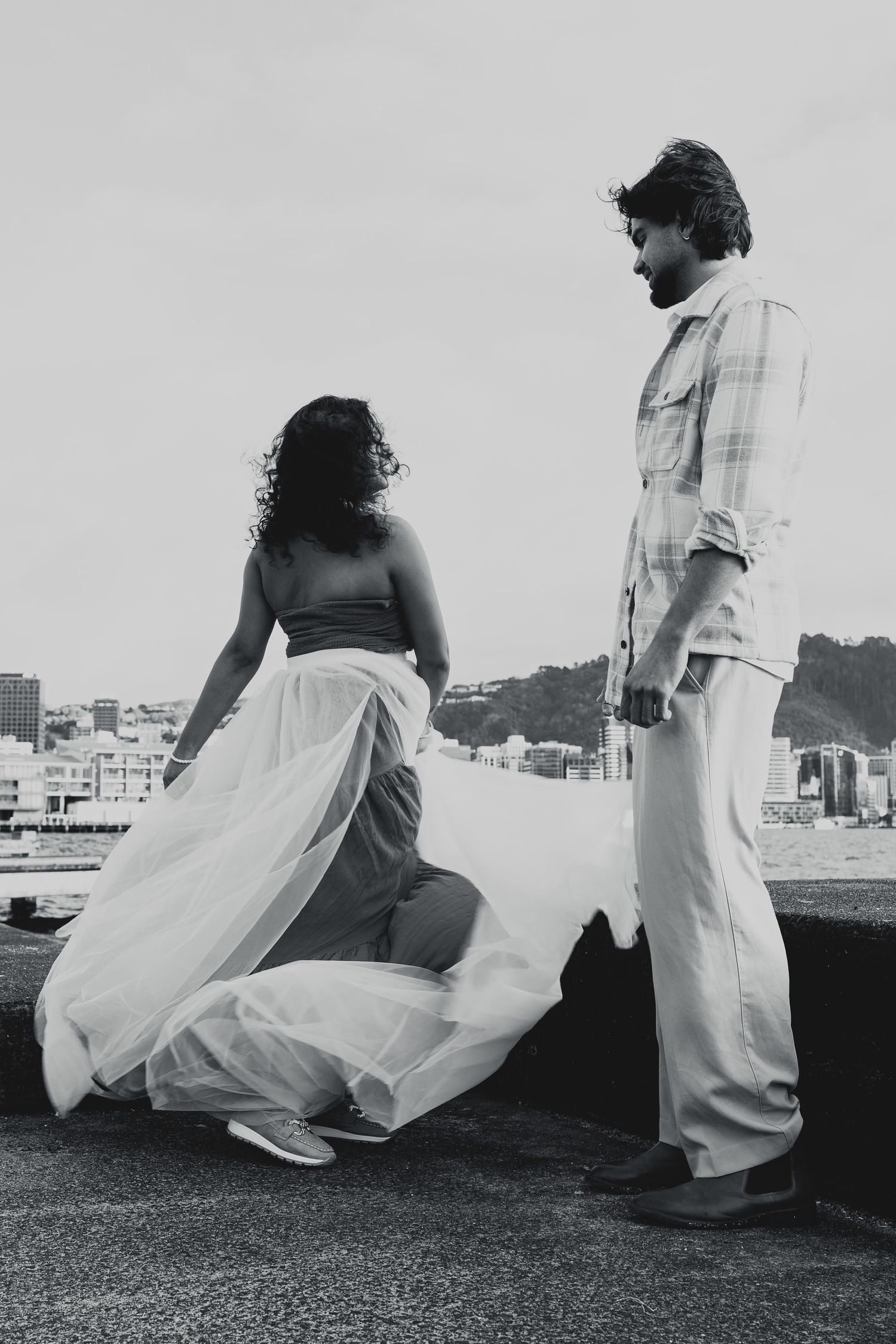 Black and white photo of a couple standing outdoors, woman in flowing skirt, man in plaid shirt, cityscape background.