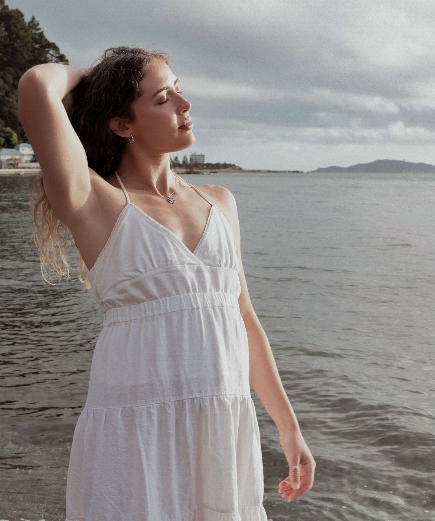 Young woman in white sundress standing by the ocean on a cloudy day.