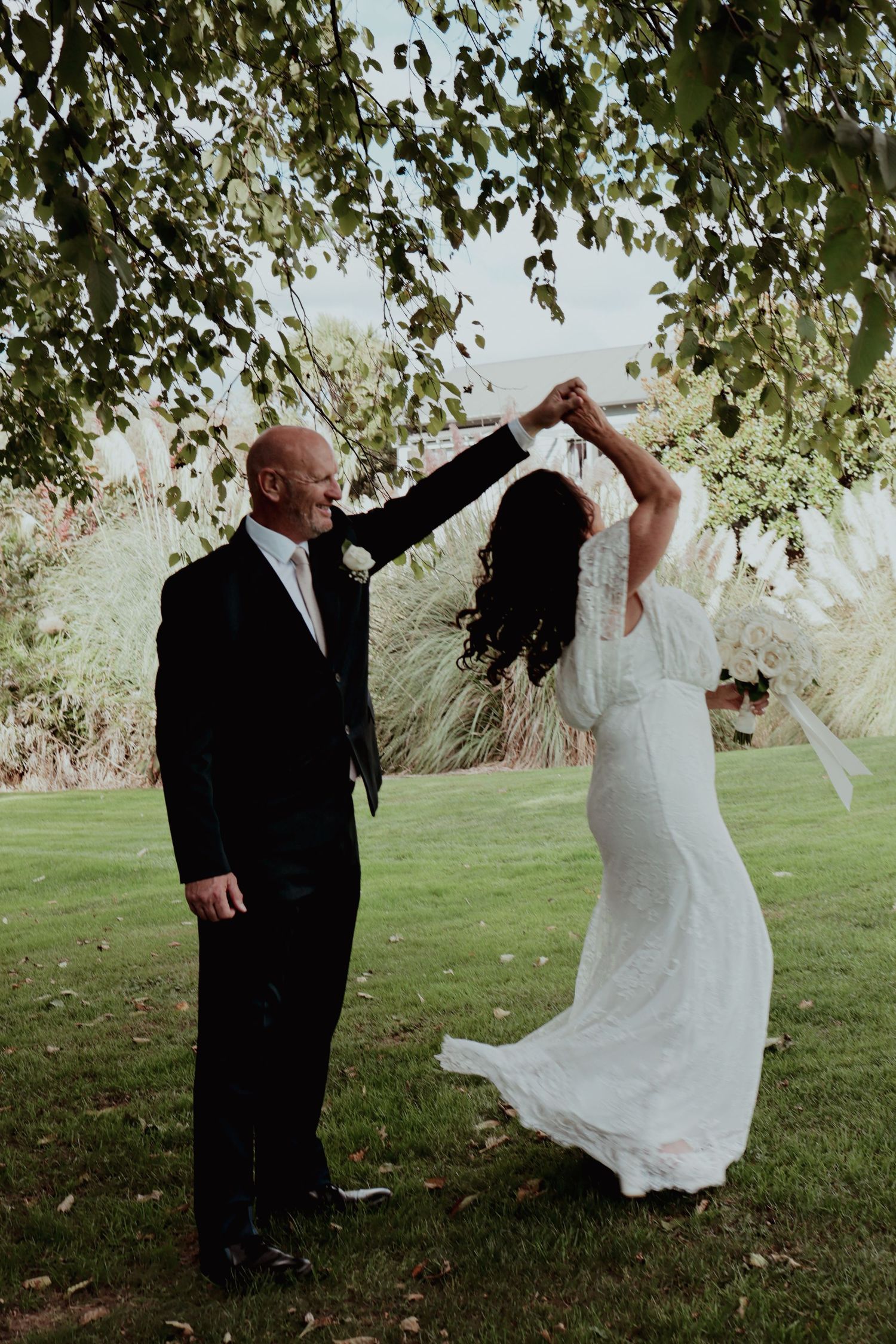 Bride and groom dancing joyfully under green trees on lush grass at outdoor wedding celebration.