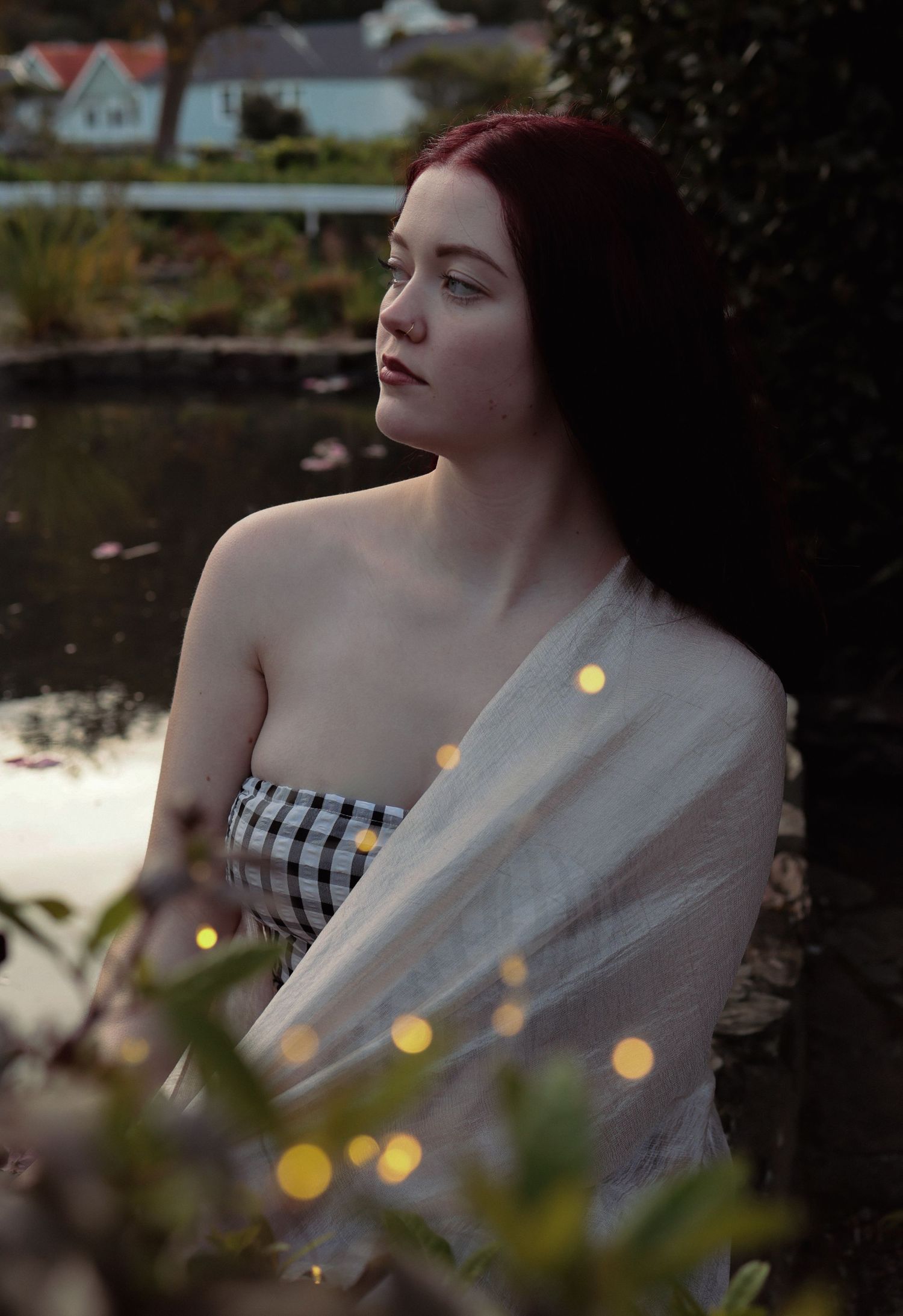 Red-haired woman in gingham top draped with sheer fabric poses near a reflective pond with bokeh fairy lights.