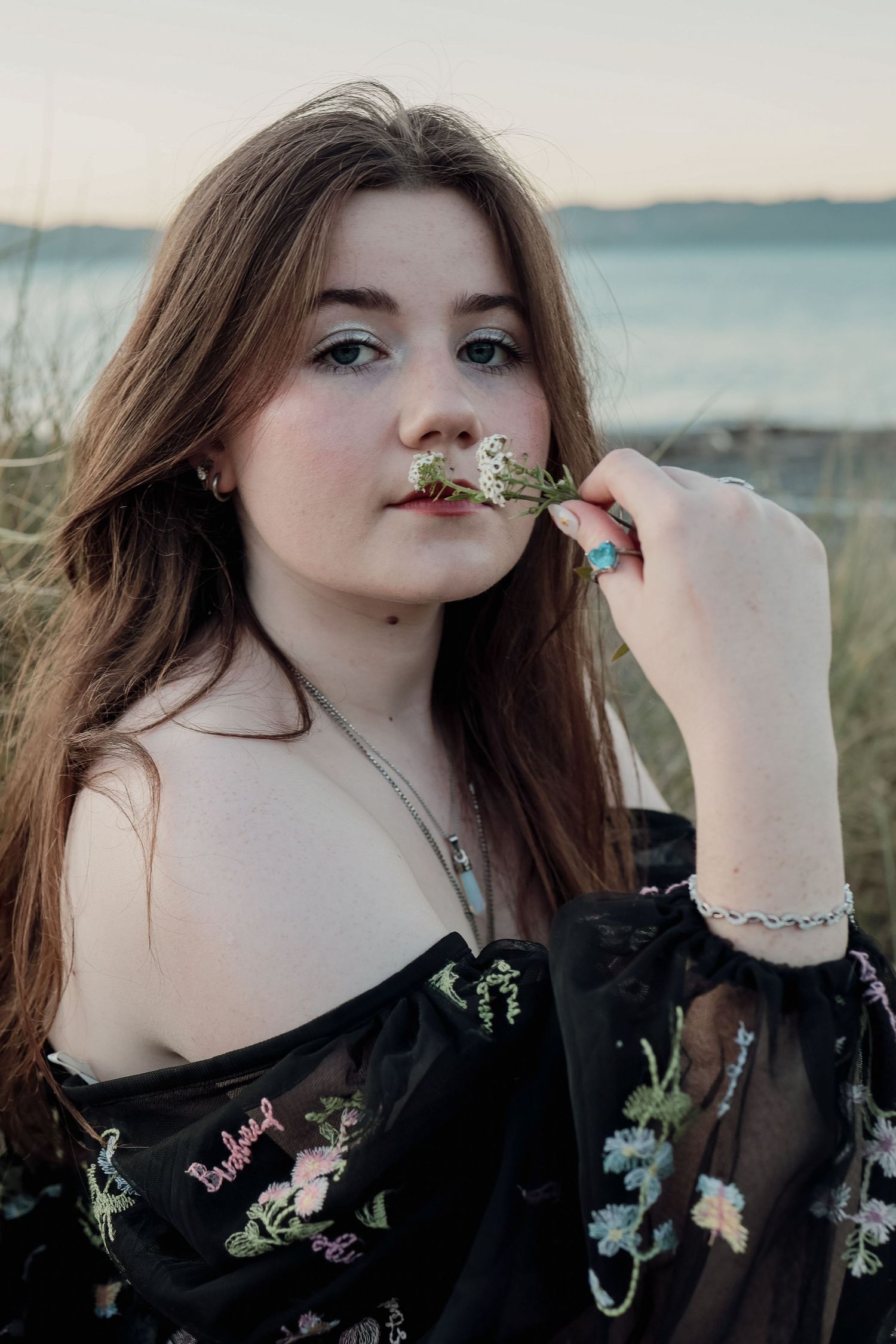 Young woman in black floral off-shoulder dress holding a flower near her lips.