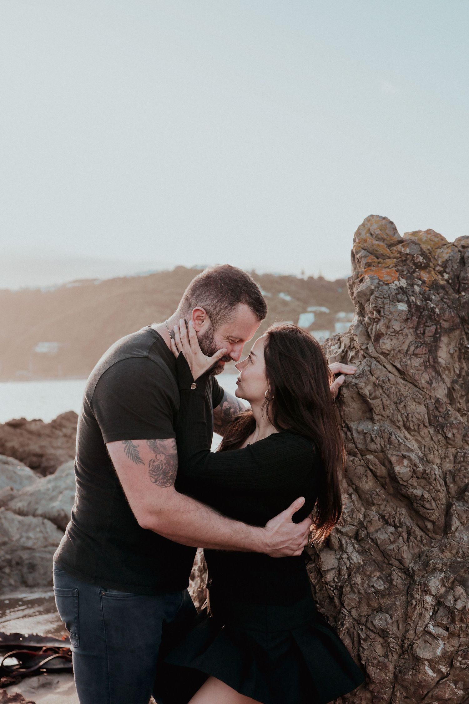 A couple shares a romantic almost-kiss moment leaning against a rocky coastal cliff at sunset.