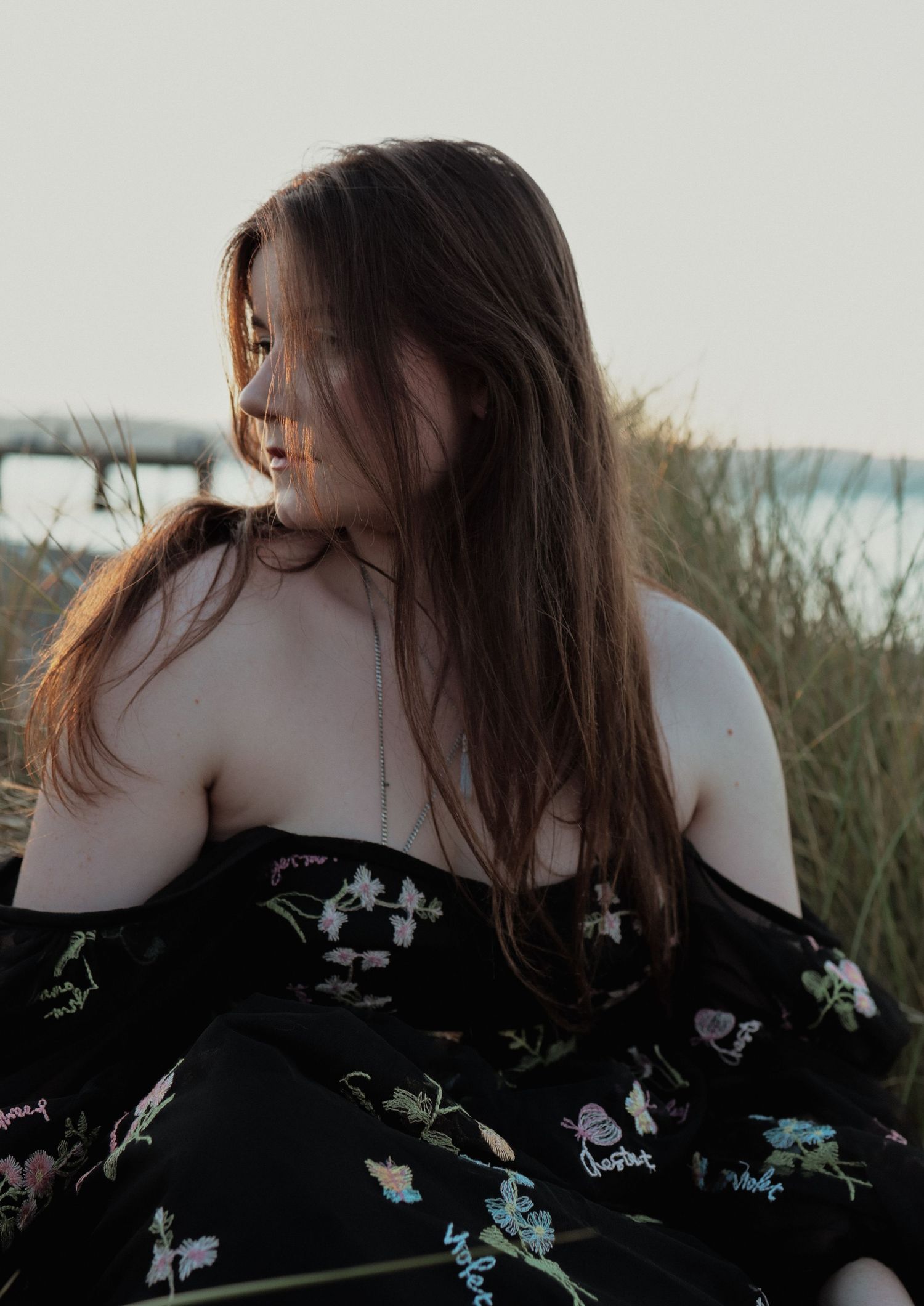 Long-haired woman in black floral off-shoulder dress sitting in coastal grass.