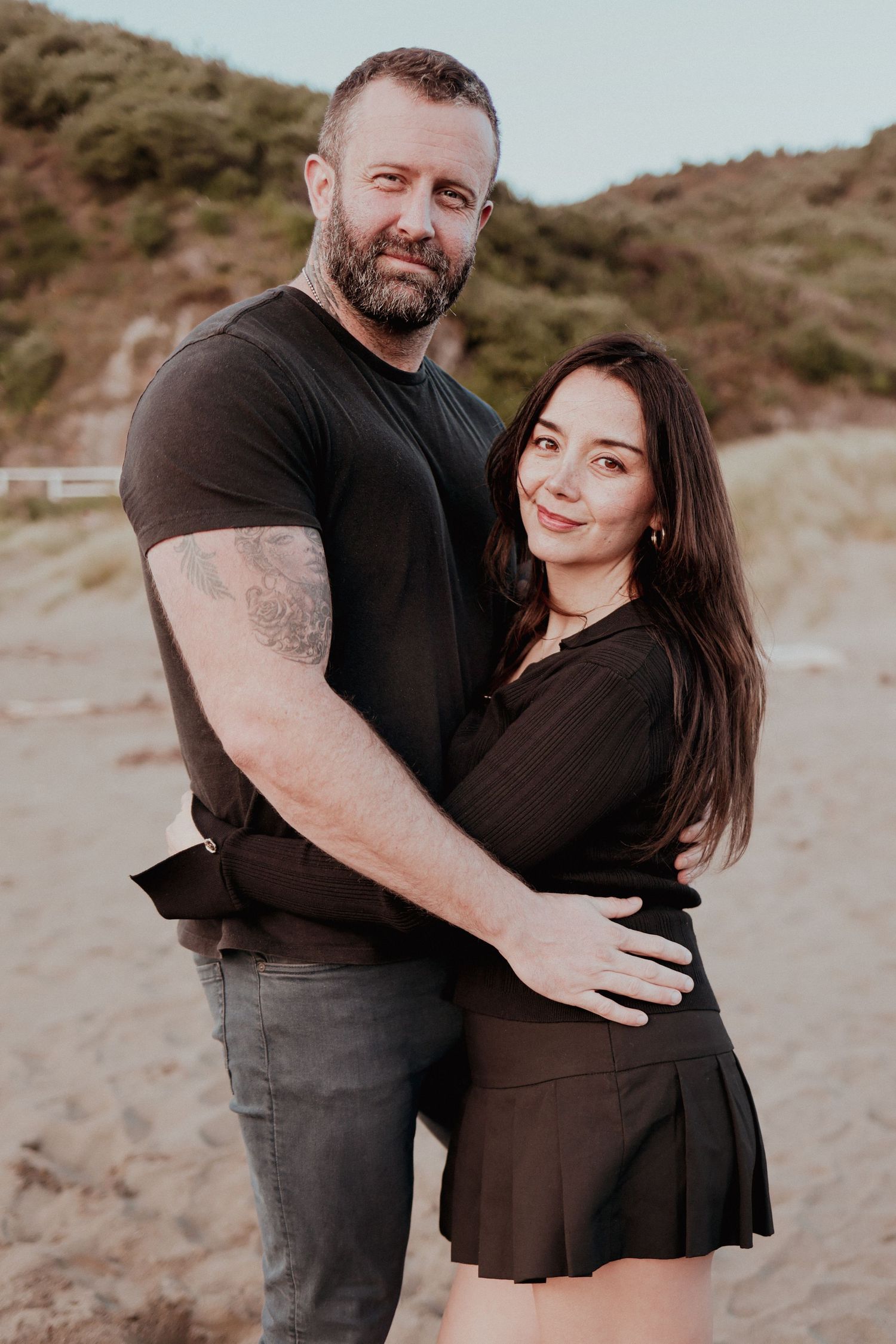 Couple embracing on sandy beach, man in black t-shirt with tattoo, woman in black outfit, hills in background.