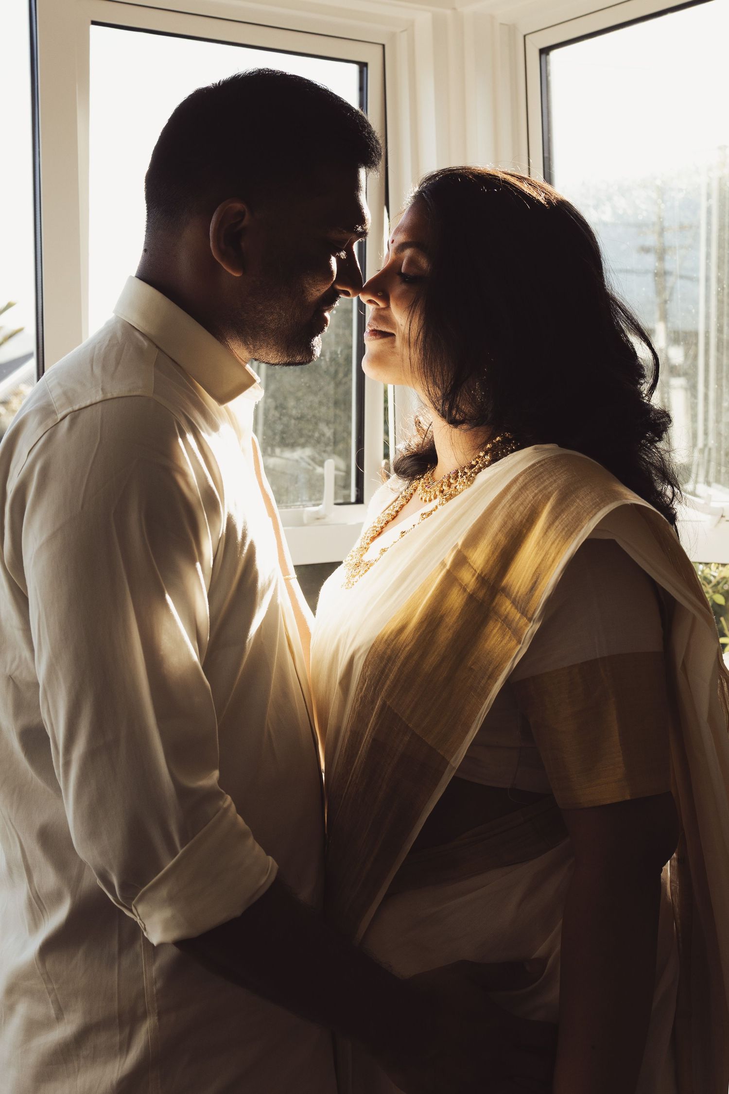 Couple in traditional Indian attire sharing a romantic moment by a sunlit window, woman in gold saree and man in white shirt.