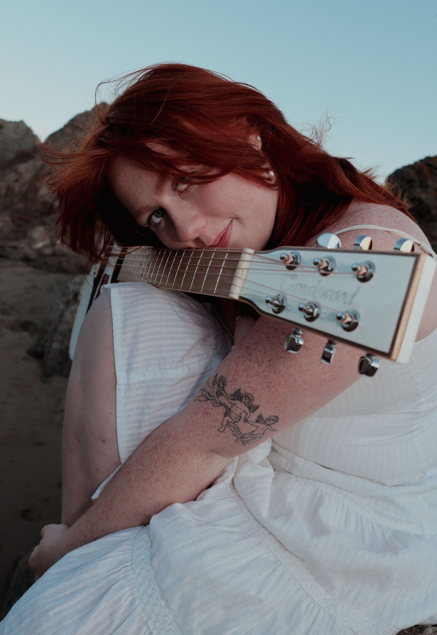 A red-haired woman in a white dress tenderly rests her cheek on an acoustic guitar outdoors.