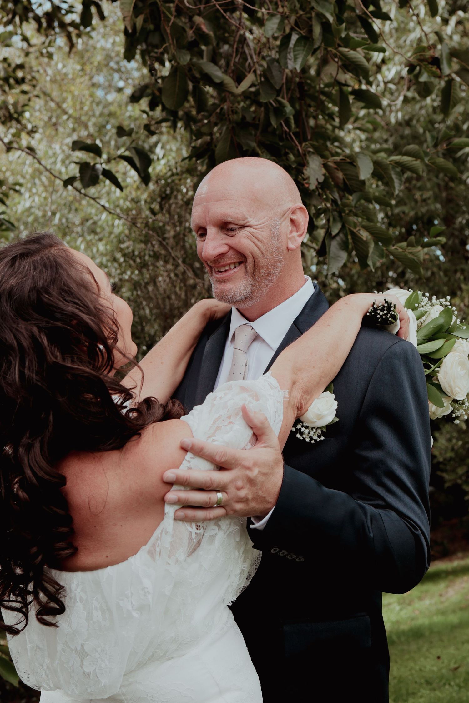 Smiling bald groom embraces bride in lace off-shoulder wedding dress in garden setting.