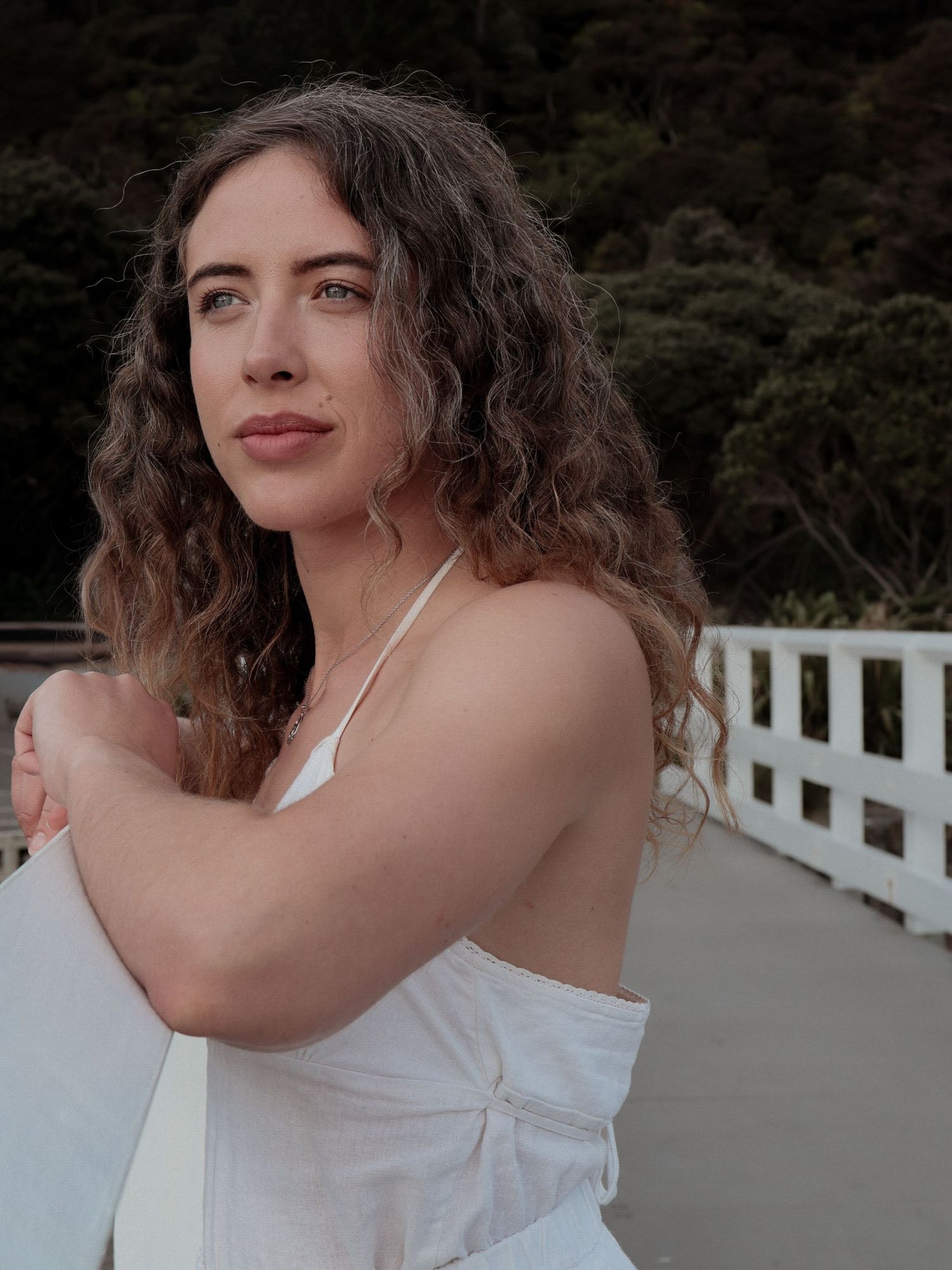Curly-haired woman in white dress hugging herself on an outdoor bridge.