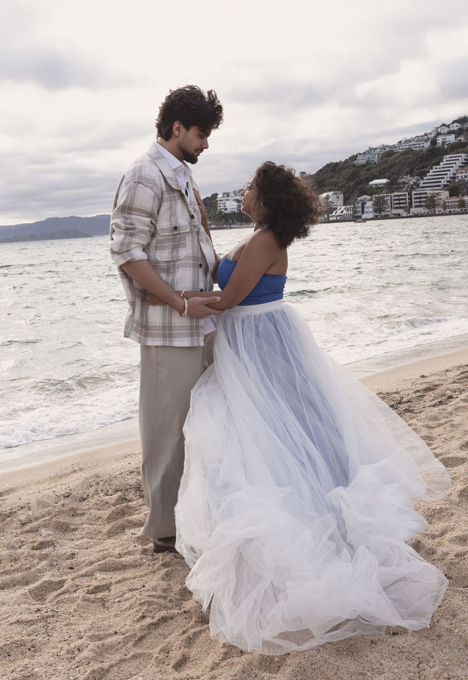 Couple in formal attire holding hands on a sandy beach with ocean waves and coastal town in the background.