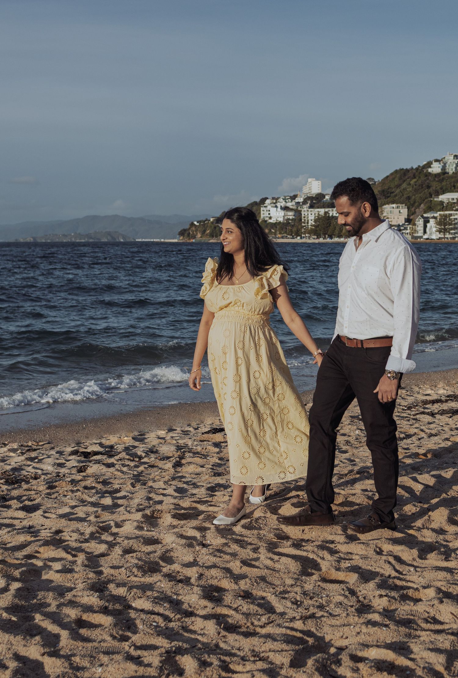 Couple walking hand in hand on a sandy beach, woman in yellow dress, man in white shirt, ocean backdrop.