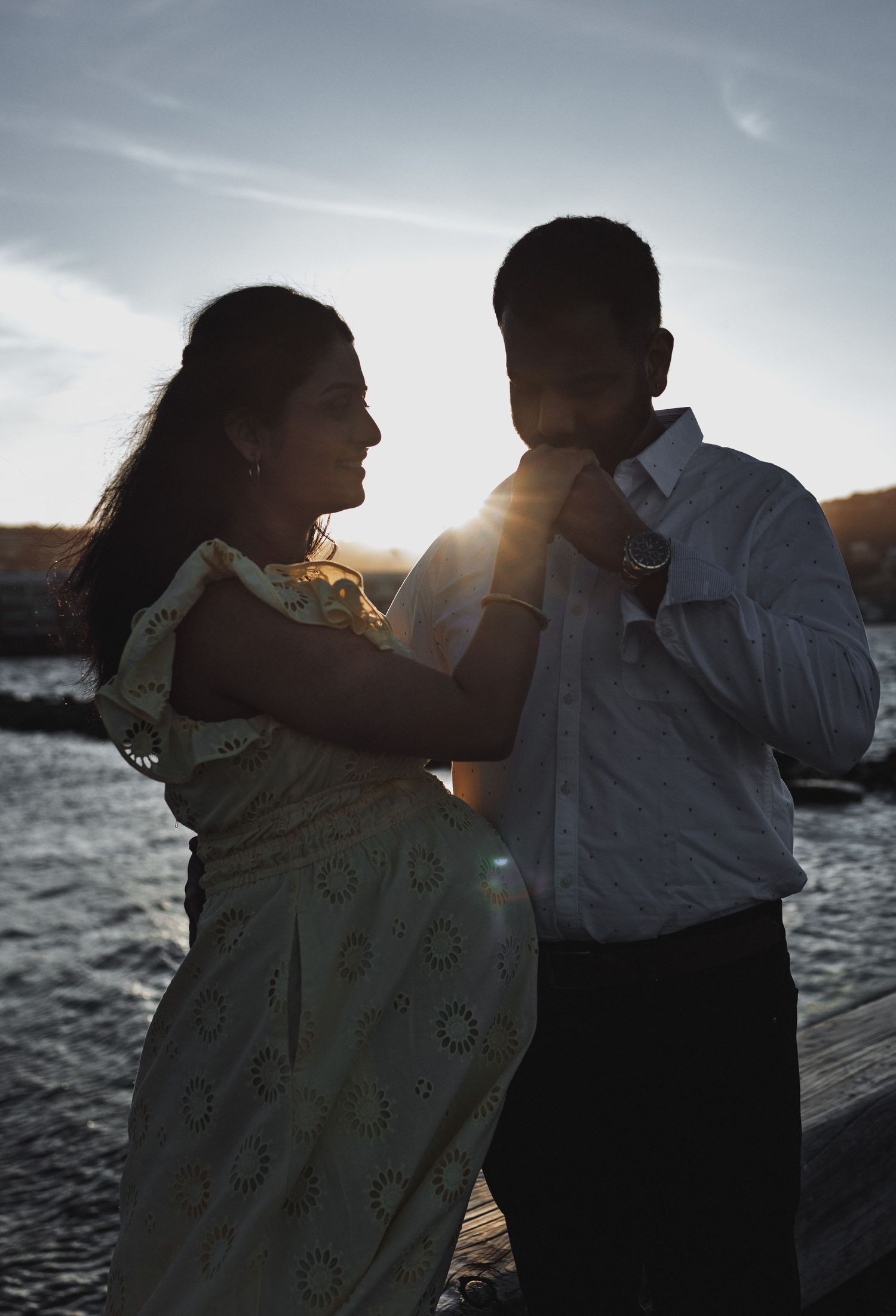Couple sharing a romantic sunset moment on a waterfront dock, woman adjusting mans tie in golden backlight.