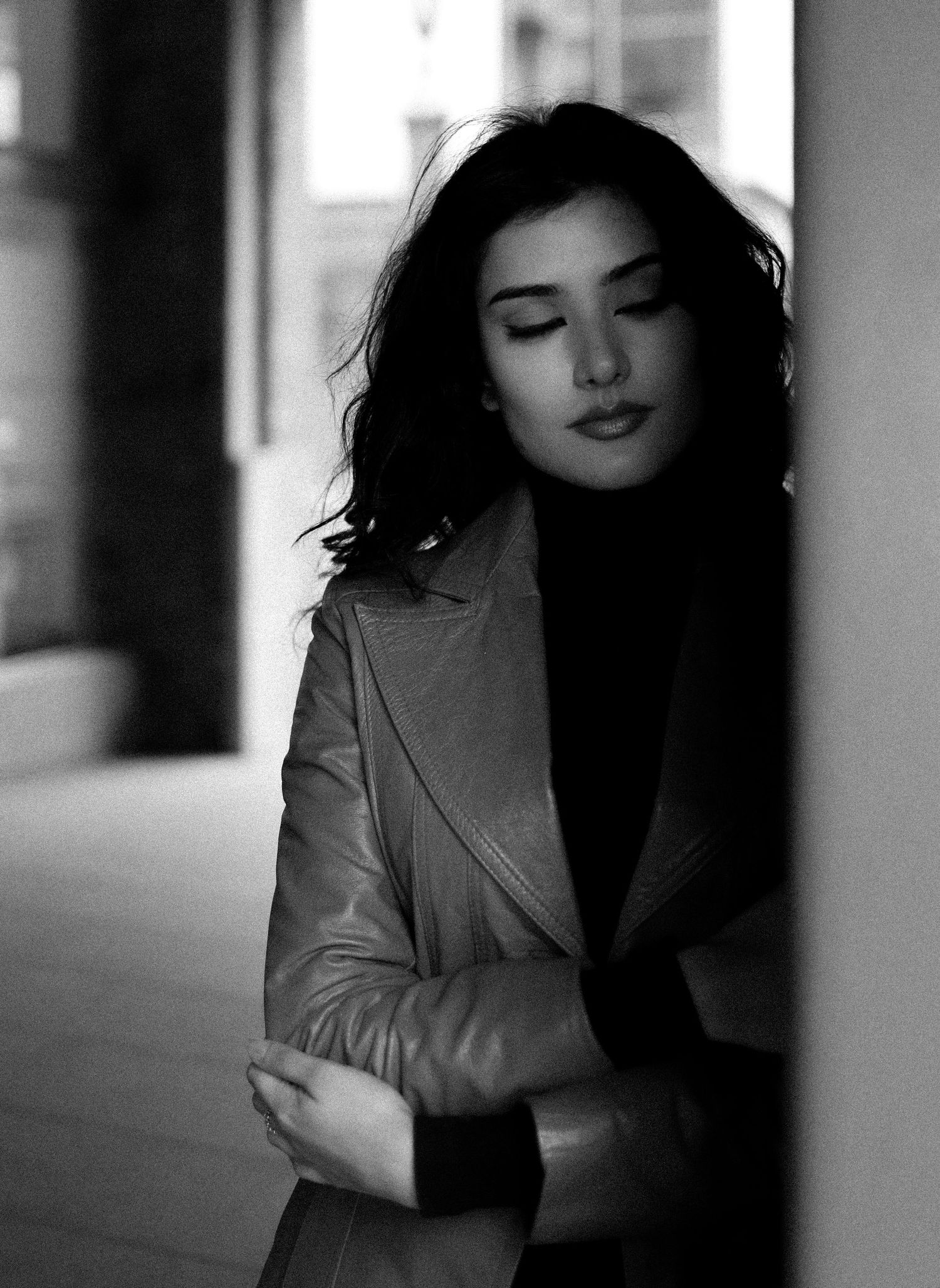 Black and white portrait of a young woman with dark wavy hair leaning against a pillar in a leather jacket.