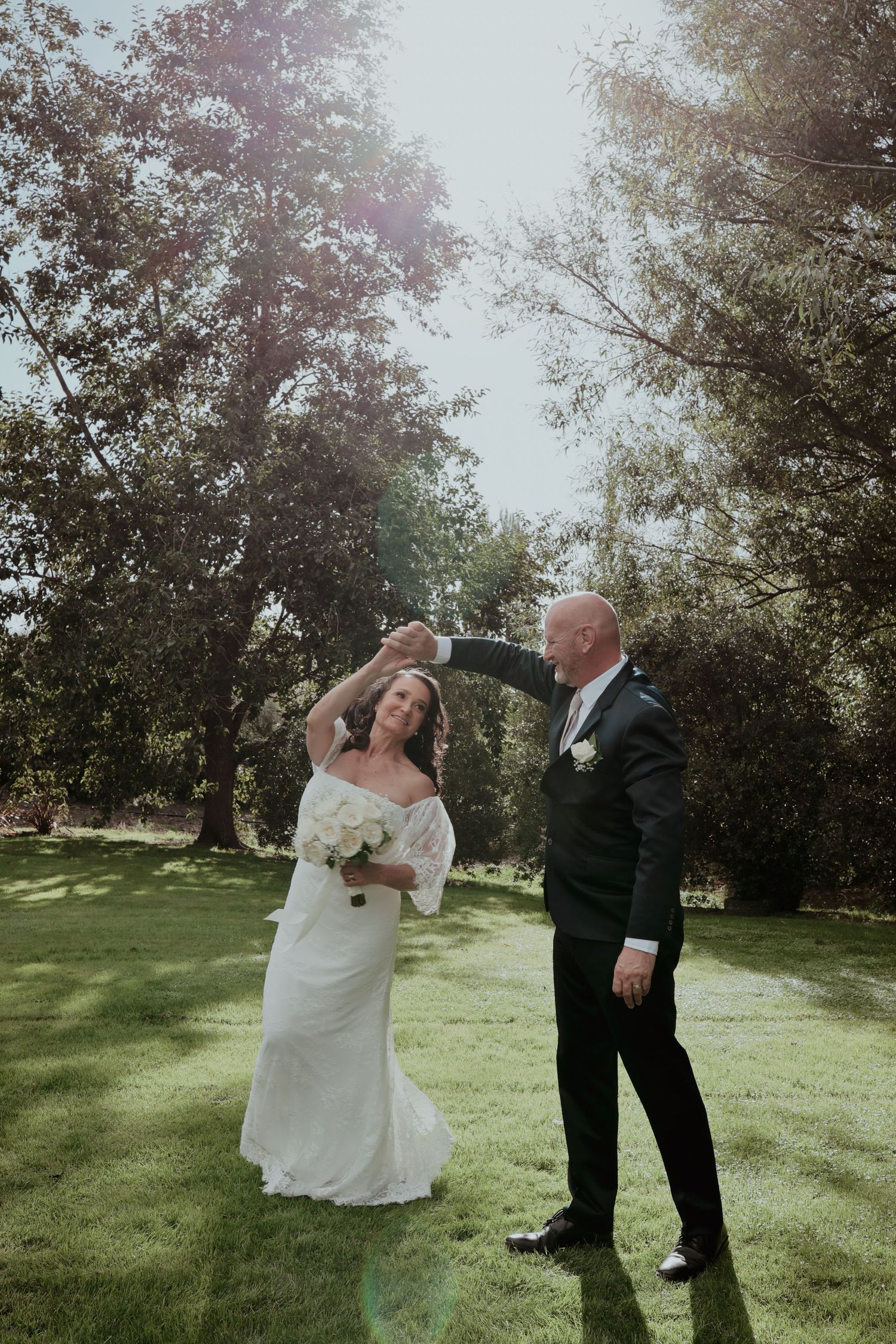 Groom twirls bride in white lace dress on sunny green lawn surrounded by trees.