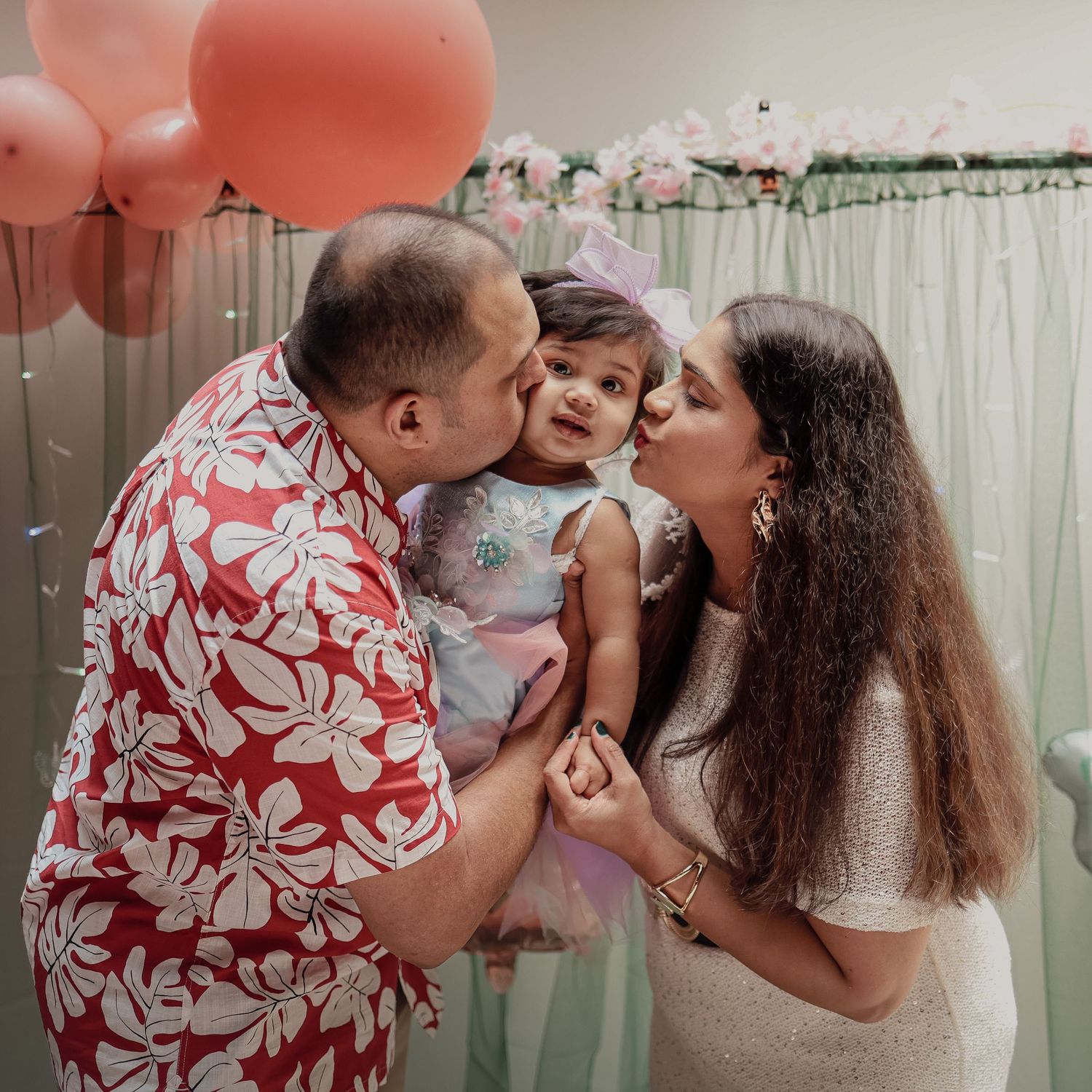 Parents in matching floral outfits kissing their baby girl on cheeks at a pink birthday party.