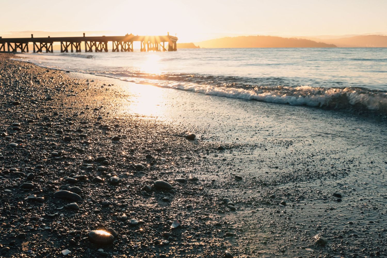 Scenic sunset beach view with gentle waves and a wooden pier in the background.