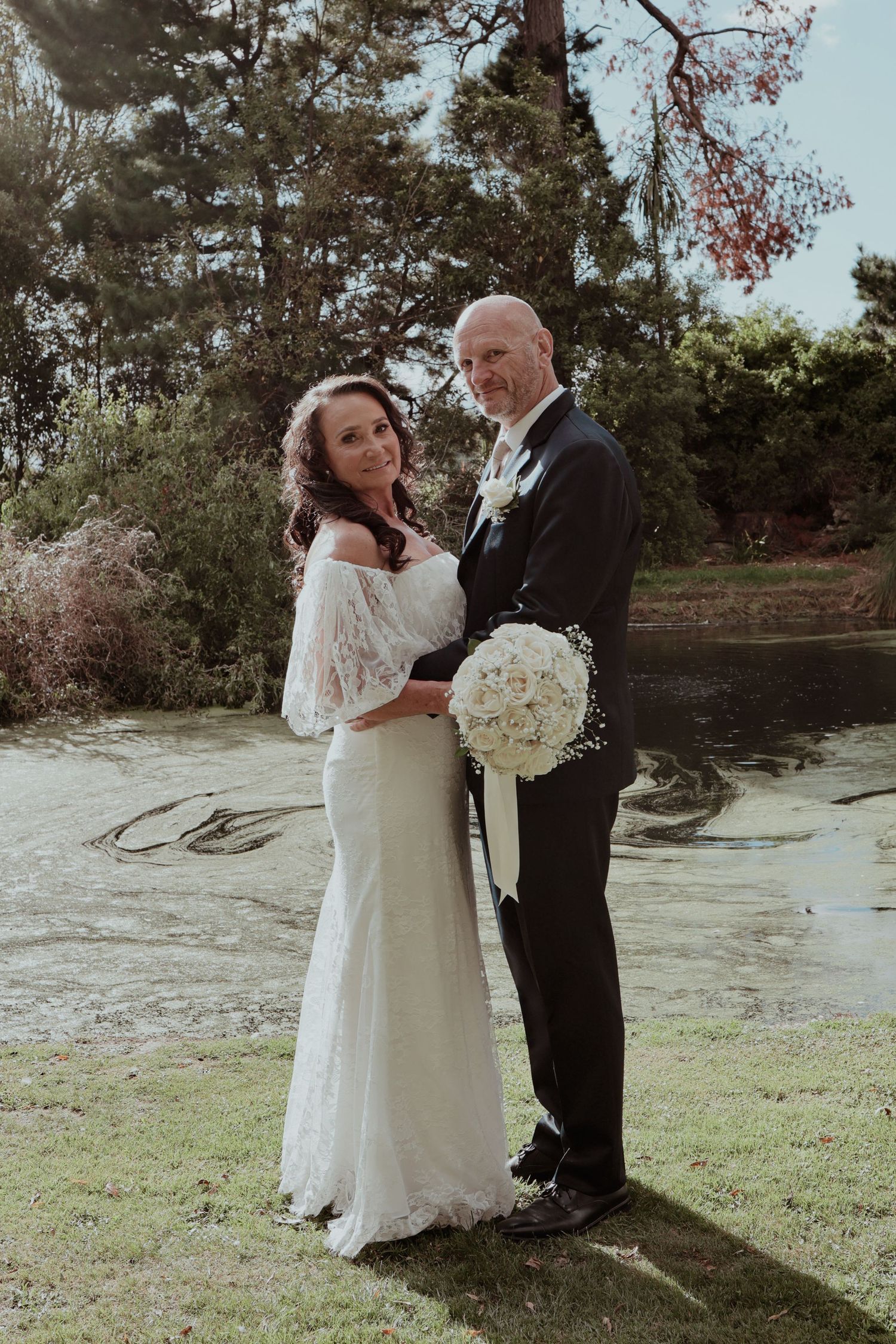 Bride and groom pose together holding white rose bouquet beside reflective pond outdoors.