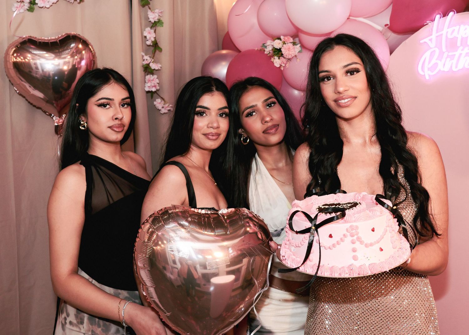 Four young women posing together at a pink-themed birthday party with heart-shaped balloons.