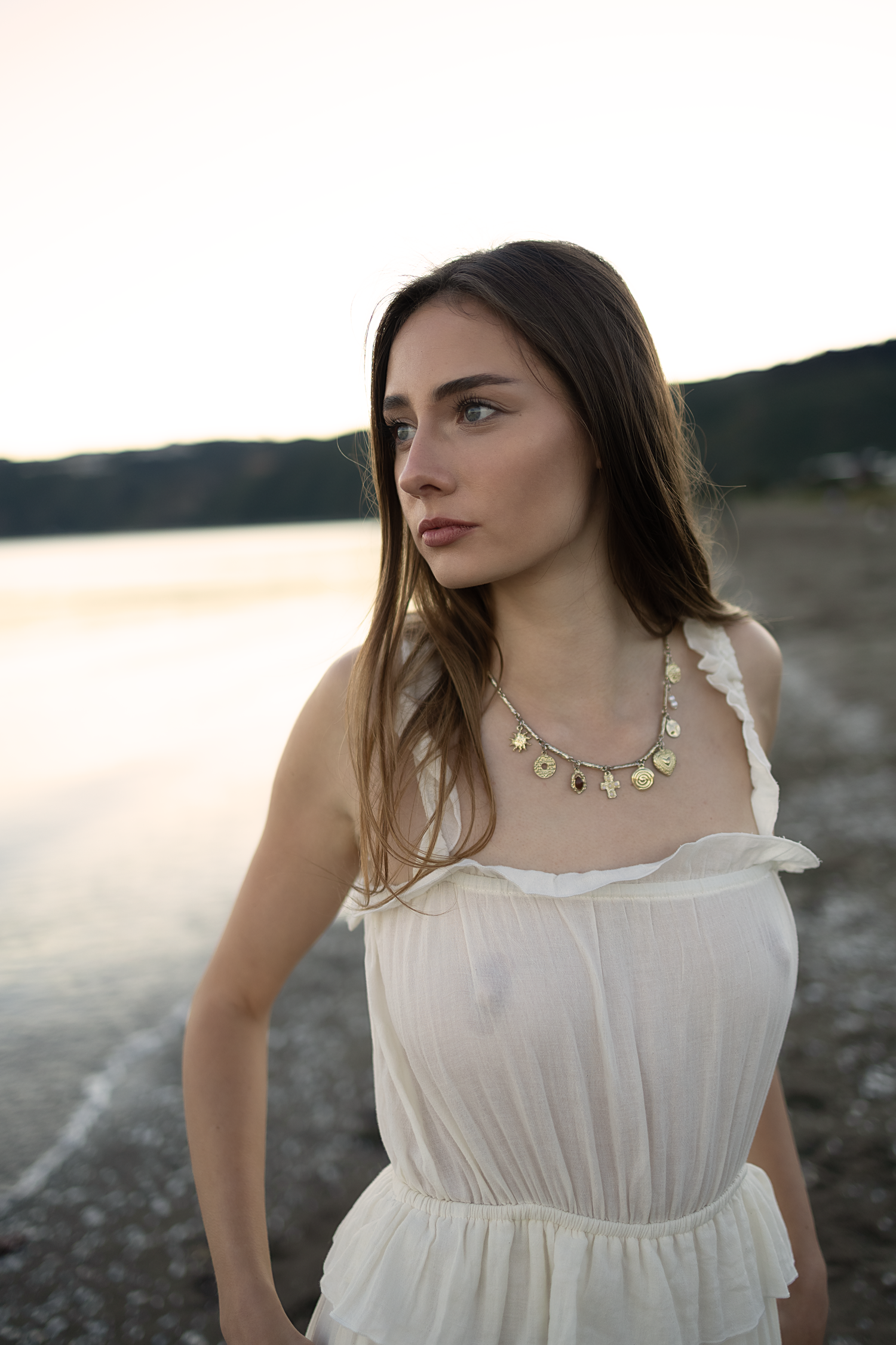 Young woman in white dress with gold coin necklace standing near a lake at sunset.