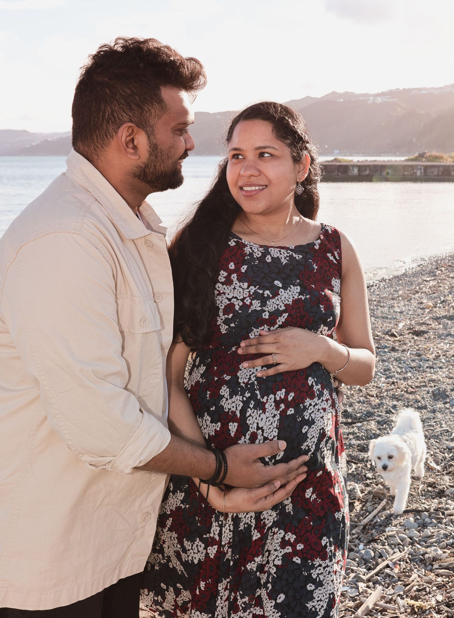 Pregnant woman in floral dress smiles at partner on rocky beach, small white dog nearby.