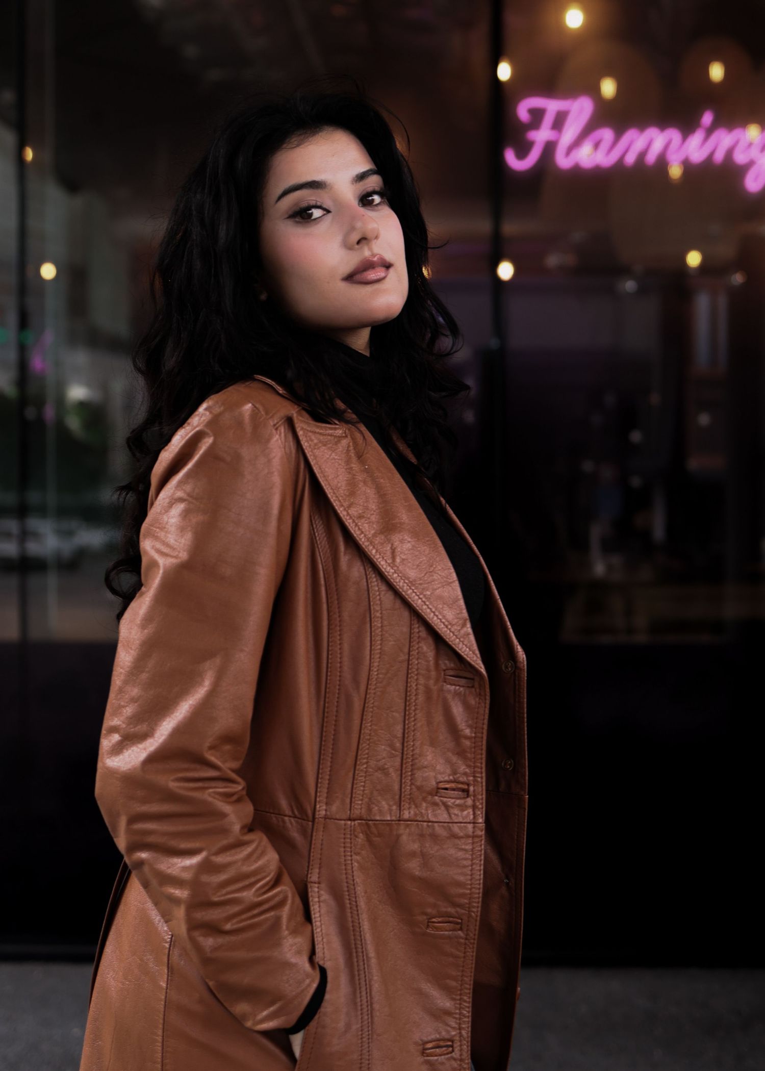 Young woman with dark curly hair in a brown leather coat posing in front of a neon Flamingo sign.