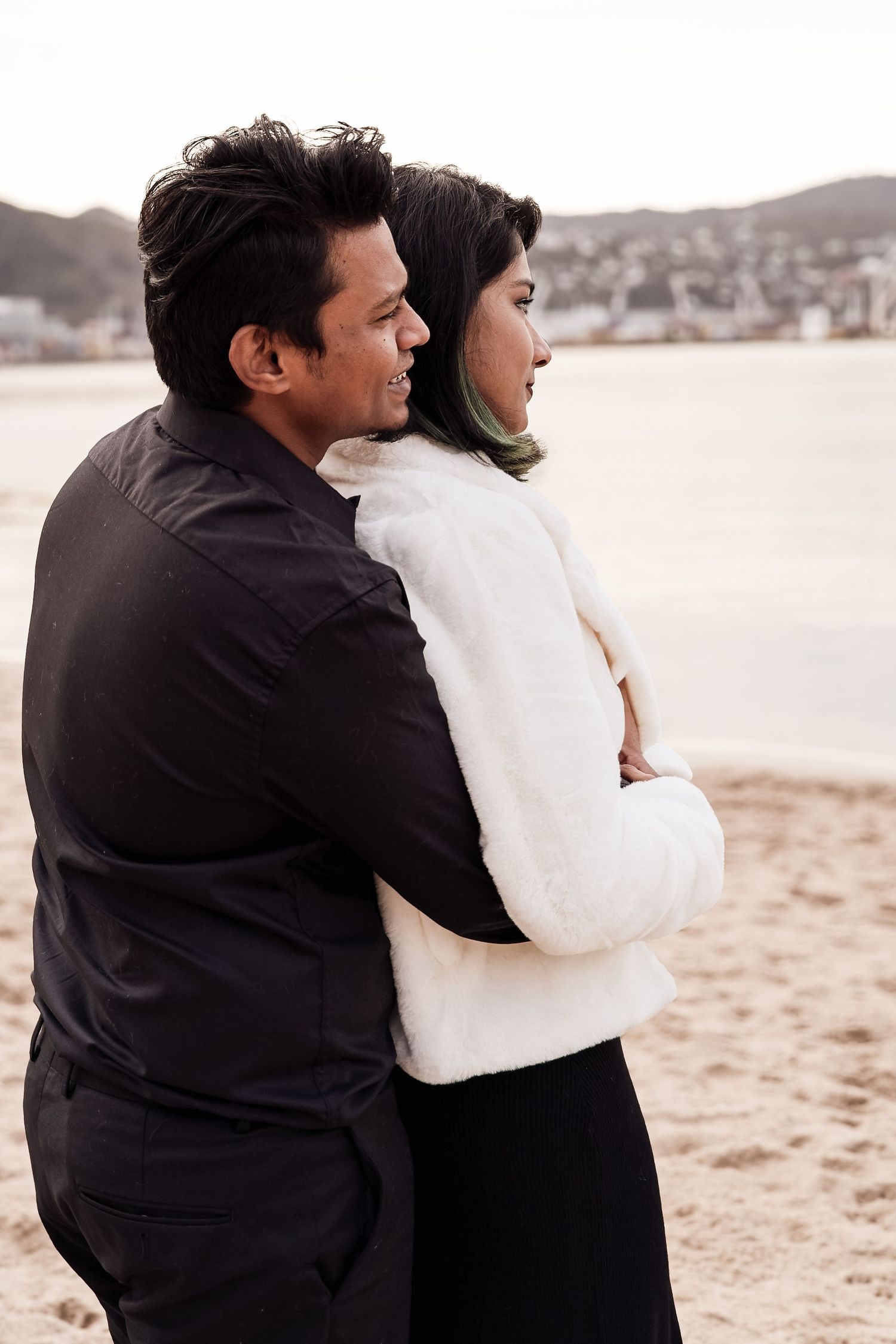 Couple embracing on a sandy beach at dusk, woman in white fur jacket, man in black shirt.
