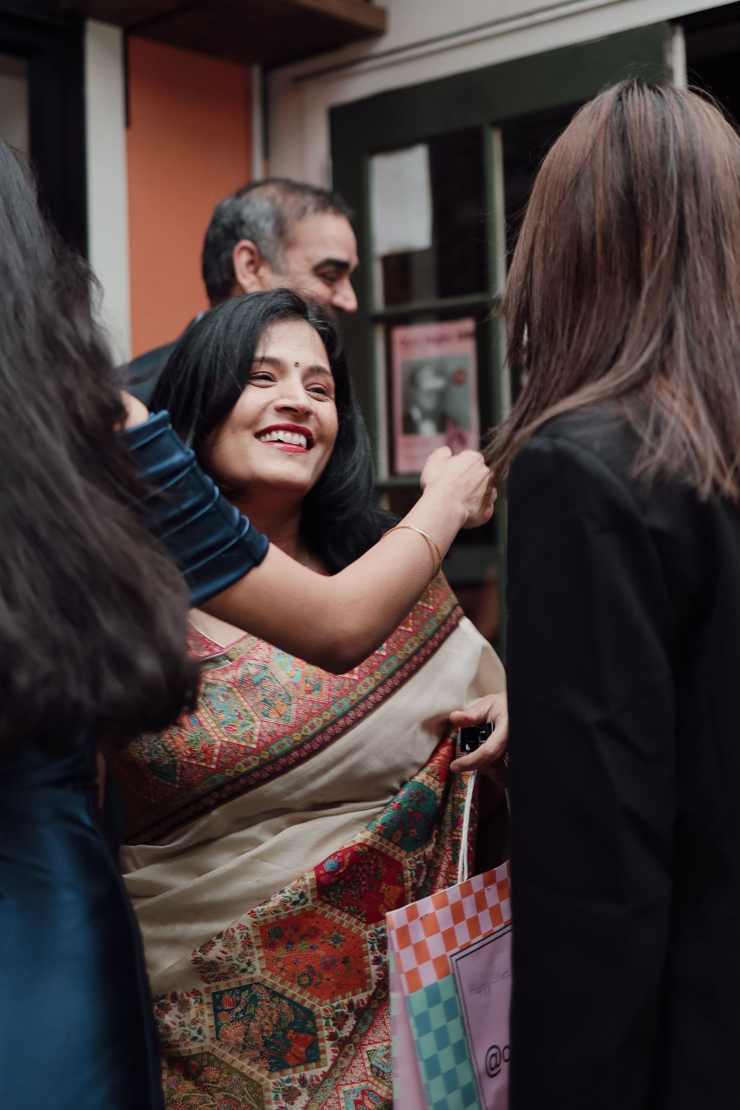 Smiling Indian woman in colorful saree hugging guests at a social gathering outdoors.