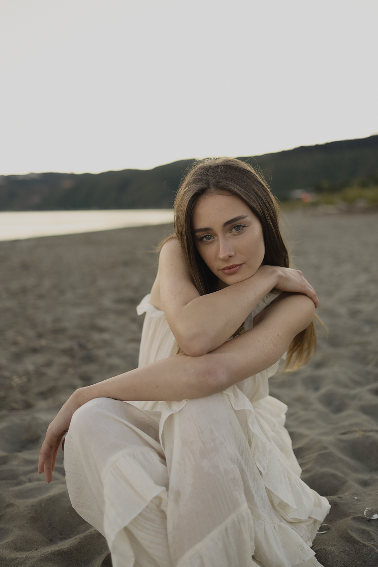 Young woman in white dress sitting on sandy beach, arms crossed, with hills and water in the background.
