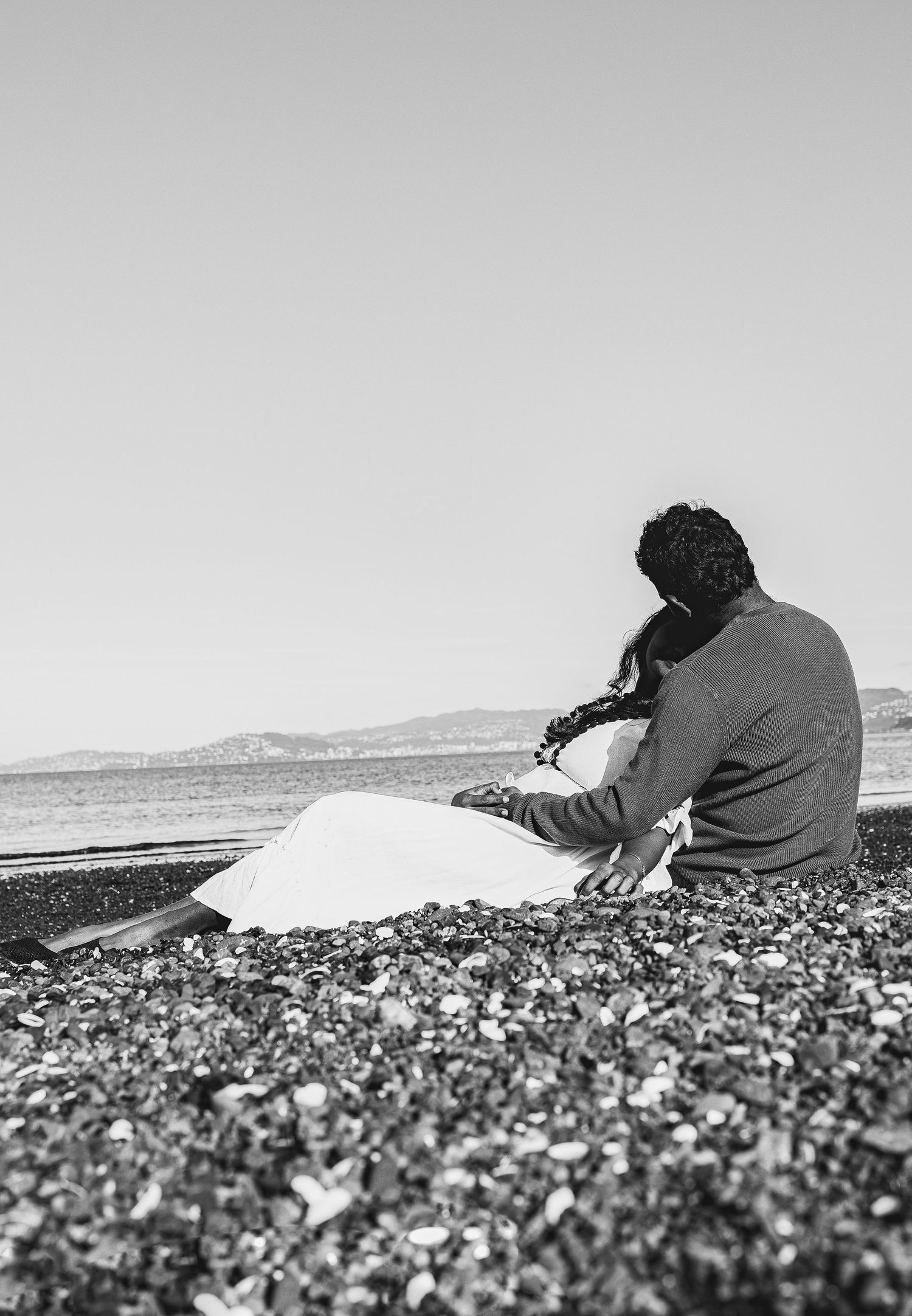 A couple sits together on a pebble beach in black and white, gazing at the calm sea with mountains in the distance.
