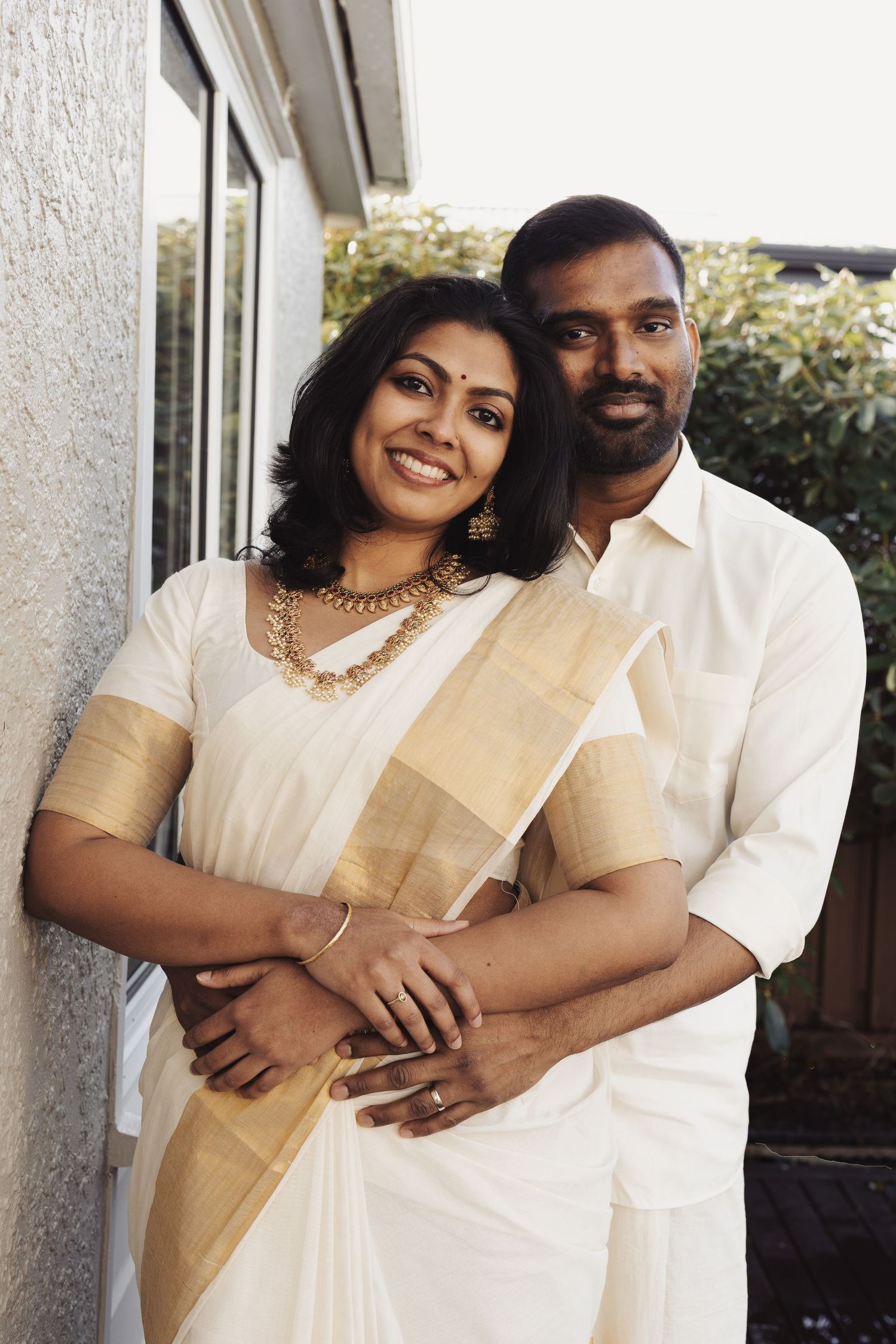 Indian couple in traditional white and gold Kerala attire smiling warmly while embracing outdoors.