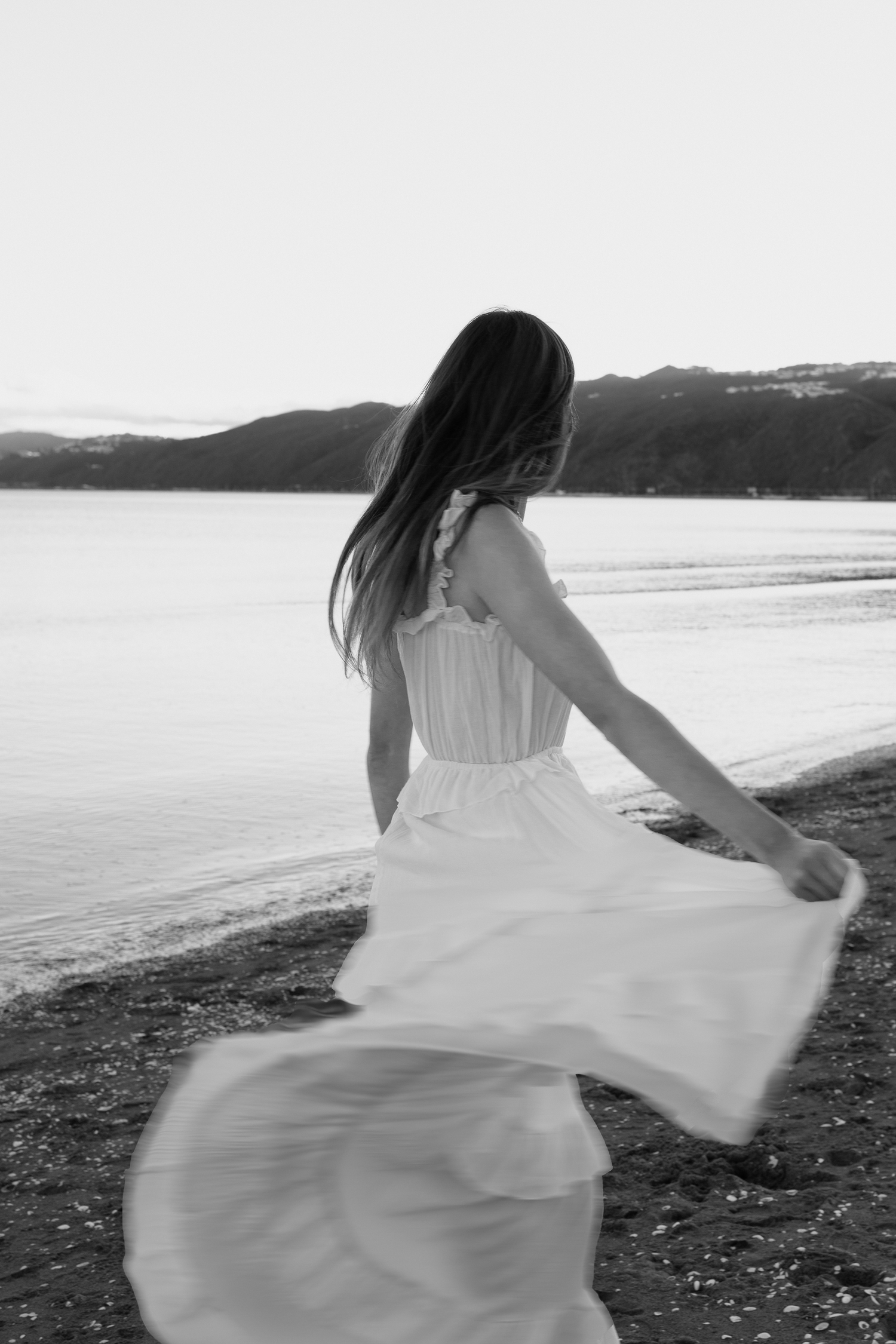 Black and white photo of a woman in a flowing white dress twirling on a rocky beach with mountains in the background.