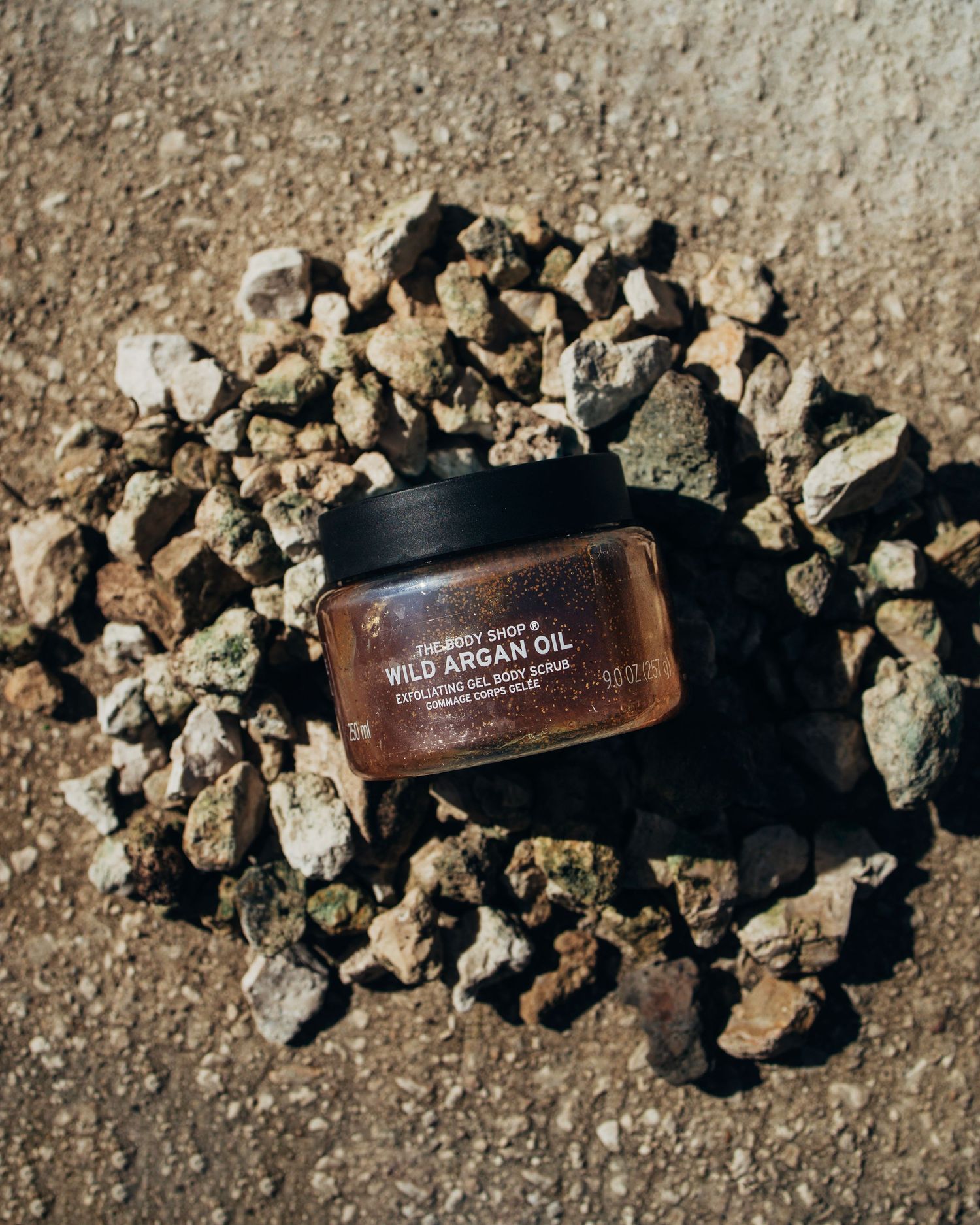 The Body Shop Wild Argan Oil exfoliating gel body scrub jar displayed on rocky ground outdoors.