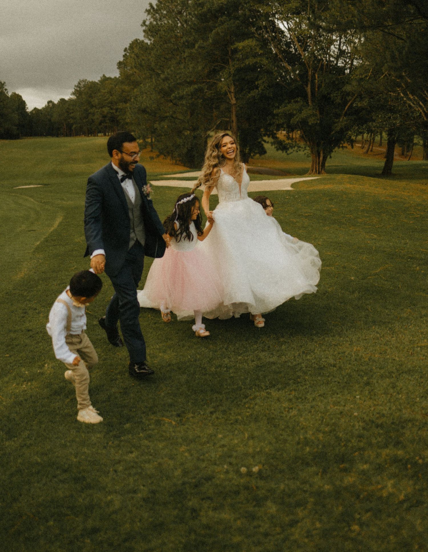 Bride in white tulle gown and groom in navy suit walk with children on green golf course at outdoor wedding.