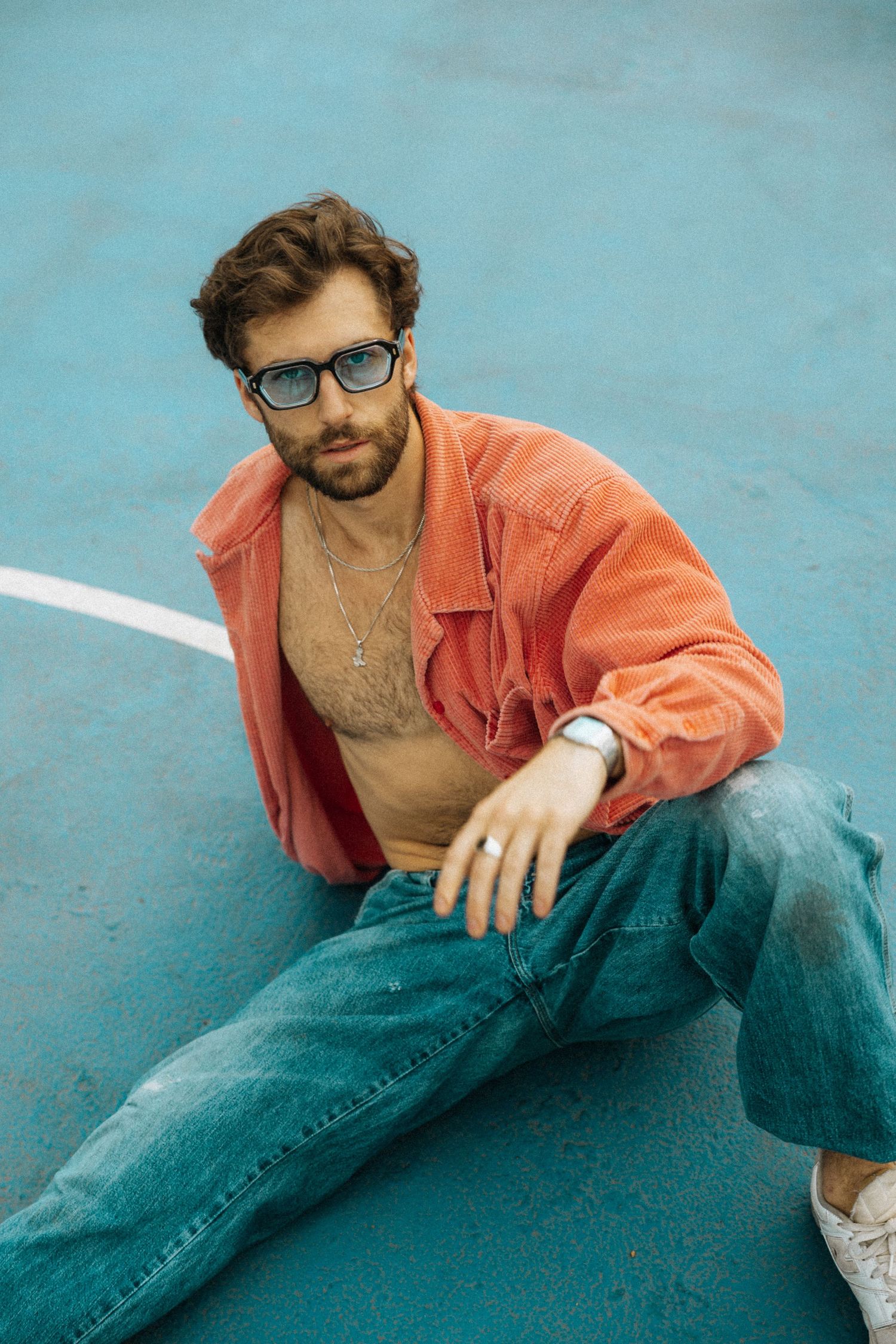 Stylish man in open coral shirt, jeans, and glasses sitting on a blue court surface with a confident pose.