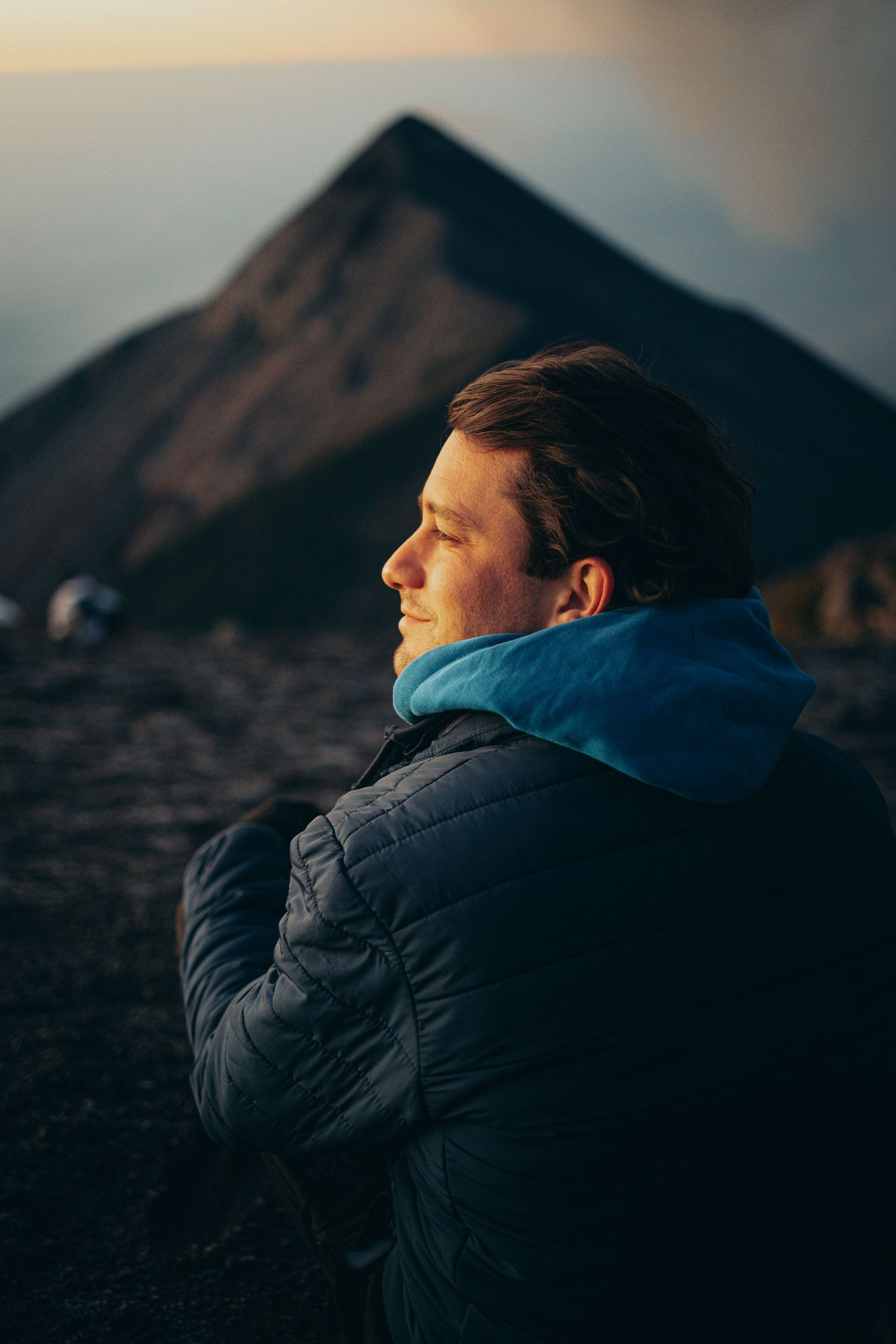 Young man in puffer jacket sitting at volcanic mountain summit at sunrise with dramatic peak in background.