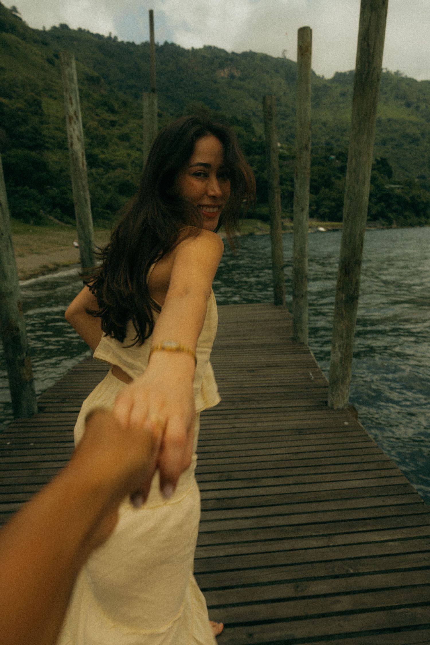 Woman in white dress leading partner by hand on wooden pier with lush green hills and water in background.