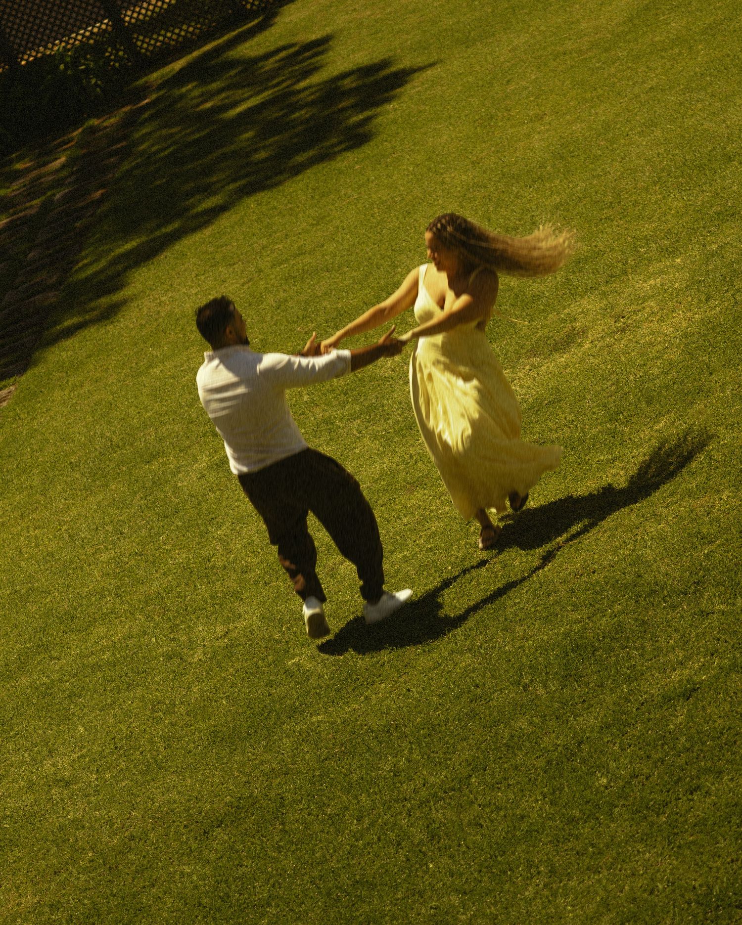 Aerial view of a couple dancing and spinning on a lush green lawn, woman in yellow dress with flowing hair.