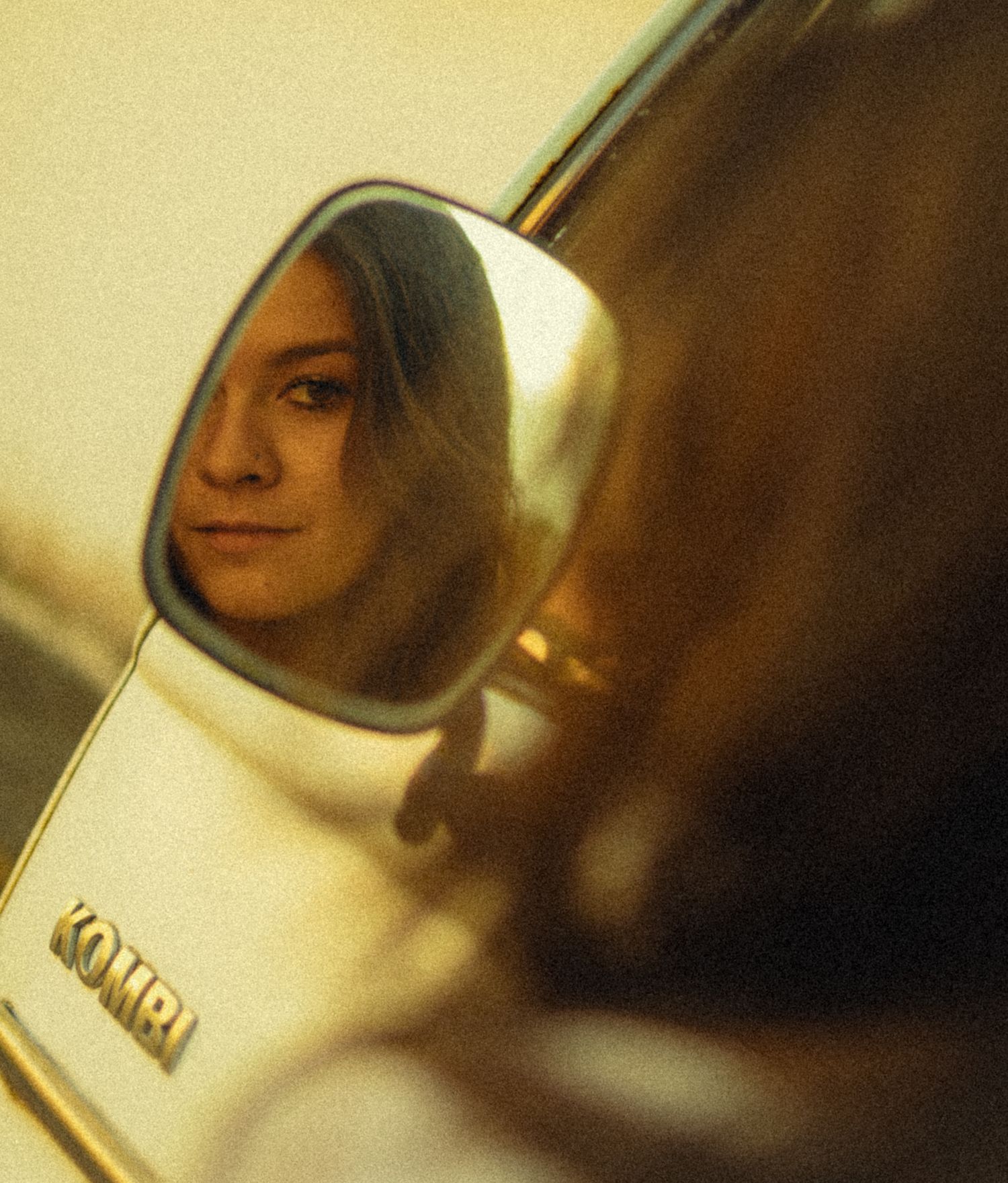 Young woman's reflection in a vintage Kombi van side mirror, bathed in warm golden light.